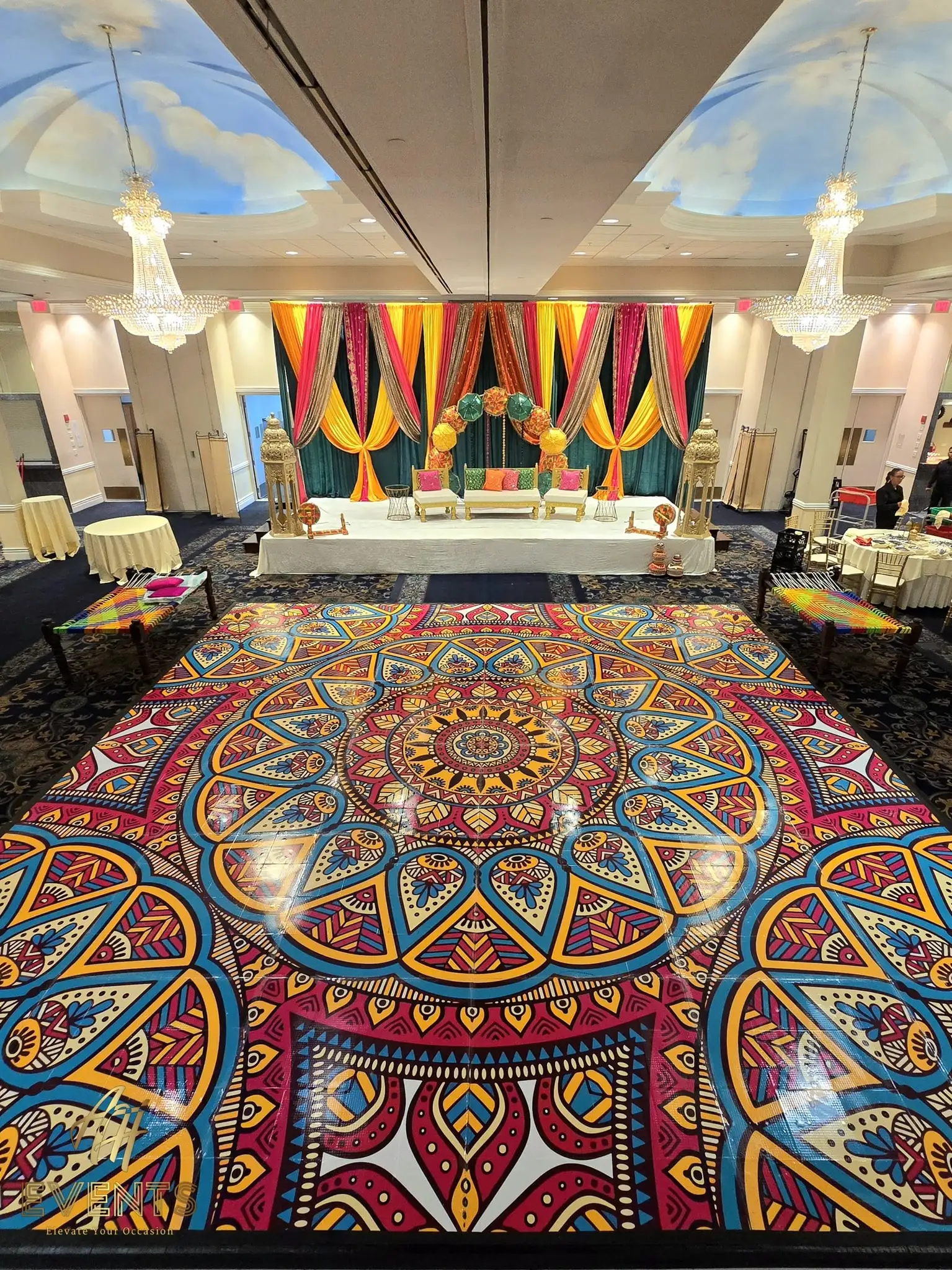 Vibrant mandala floor beneath chandeliers and a cloud-painted ceiling, facing a striped-drape stage