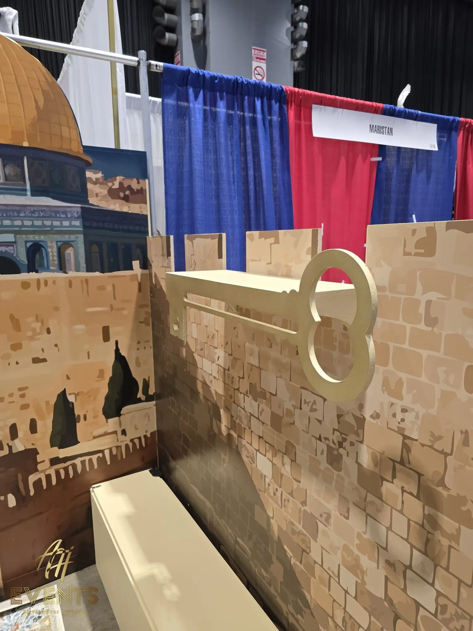 Large gold key mounted on a brick-print booth wall, with the Dome of the Rock mural and neighboring booths