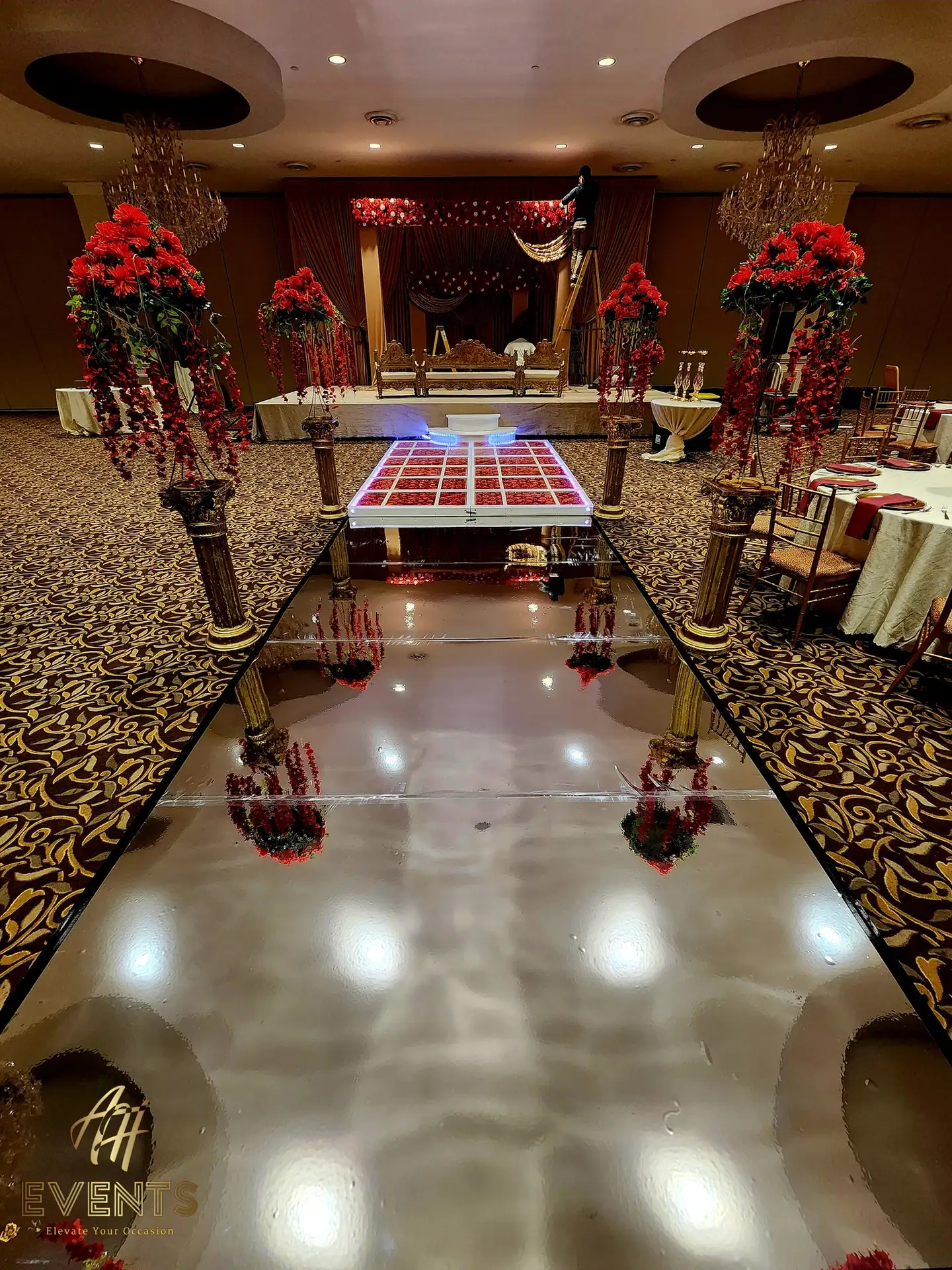 Reflective aisle leading to an LED-lit red-rose glass platform, framed by gold pillars with tall red floral arrangements
