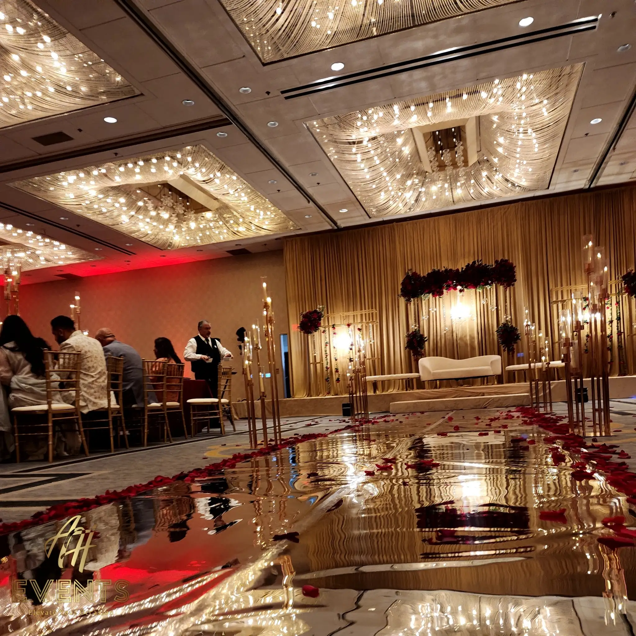 Reflective gold mirror vinyl aisle lined with gold candelabras and red petals, guests seated at tables alongside