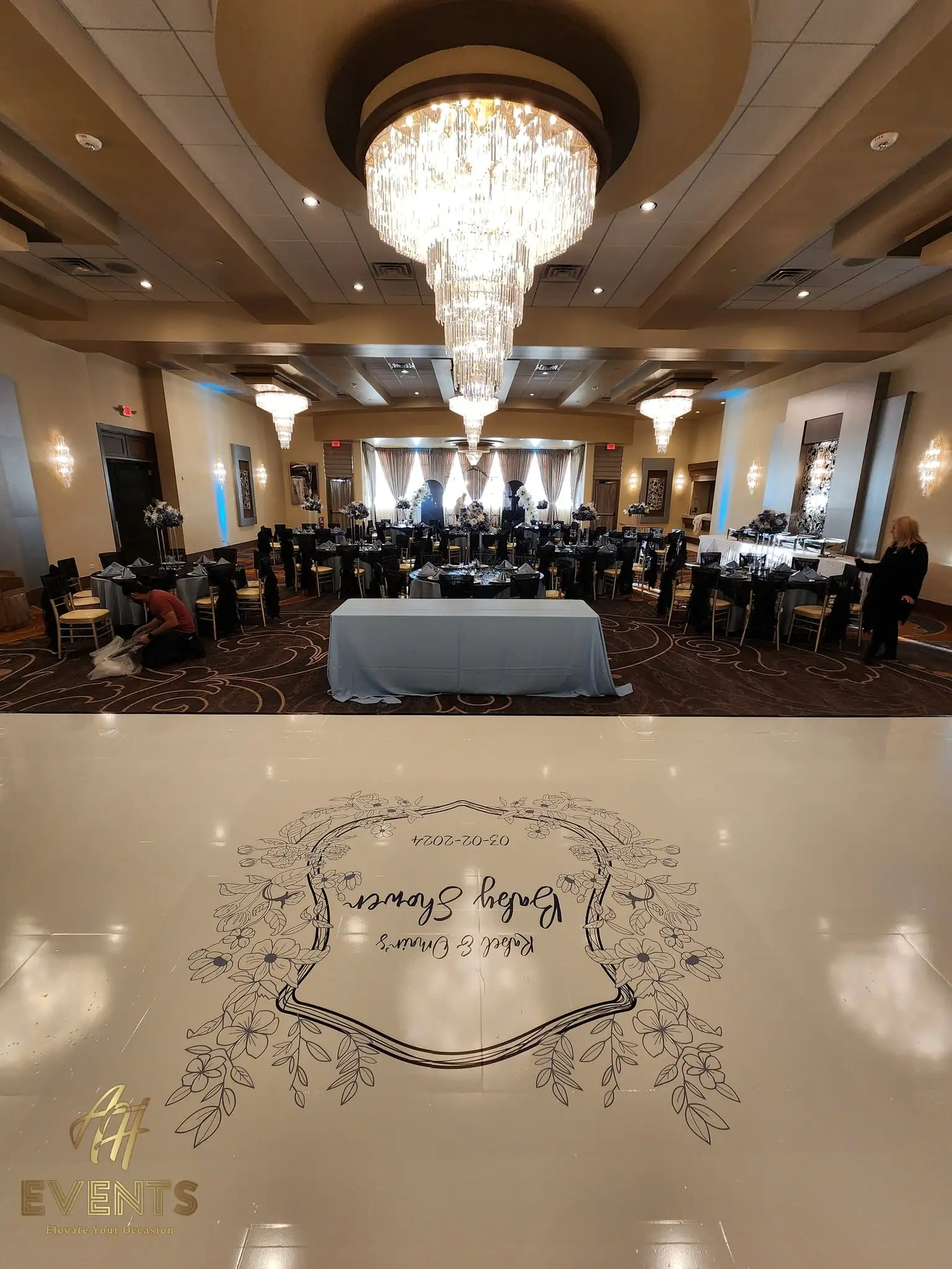 Ballroom with a crystal chandelier, the cream baby shower decal floor, a head table and an installer at work
