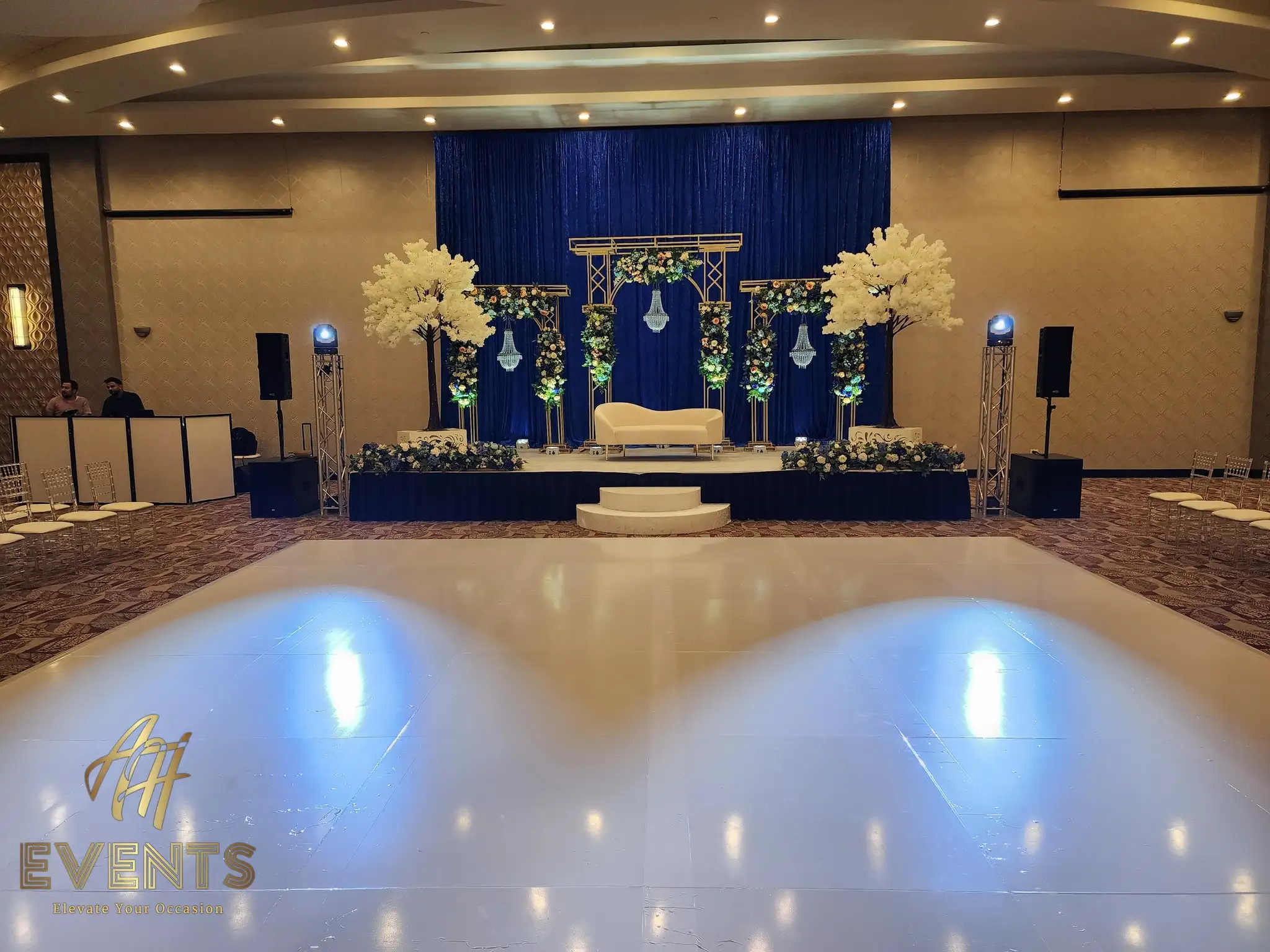 Glossy white dance floor before a navy-draped stage with white blossom trees, a floral mandap and a cream sofa