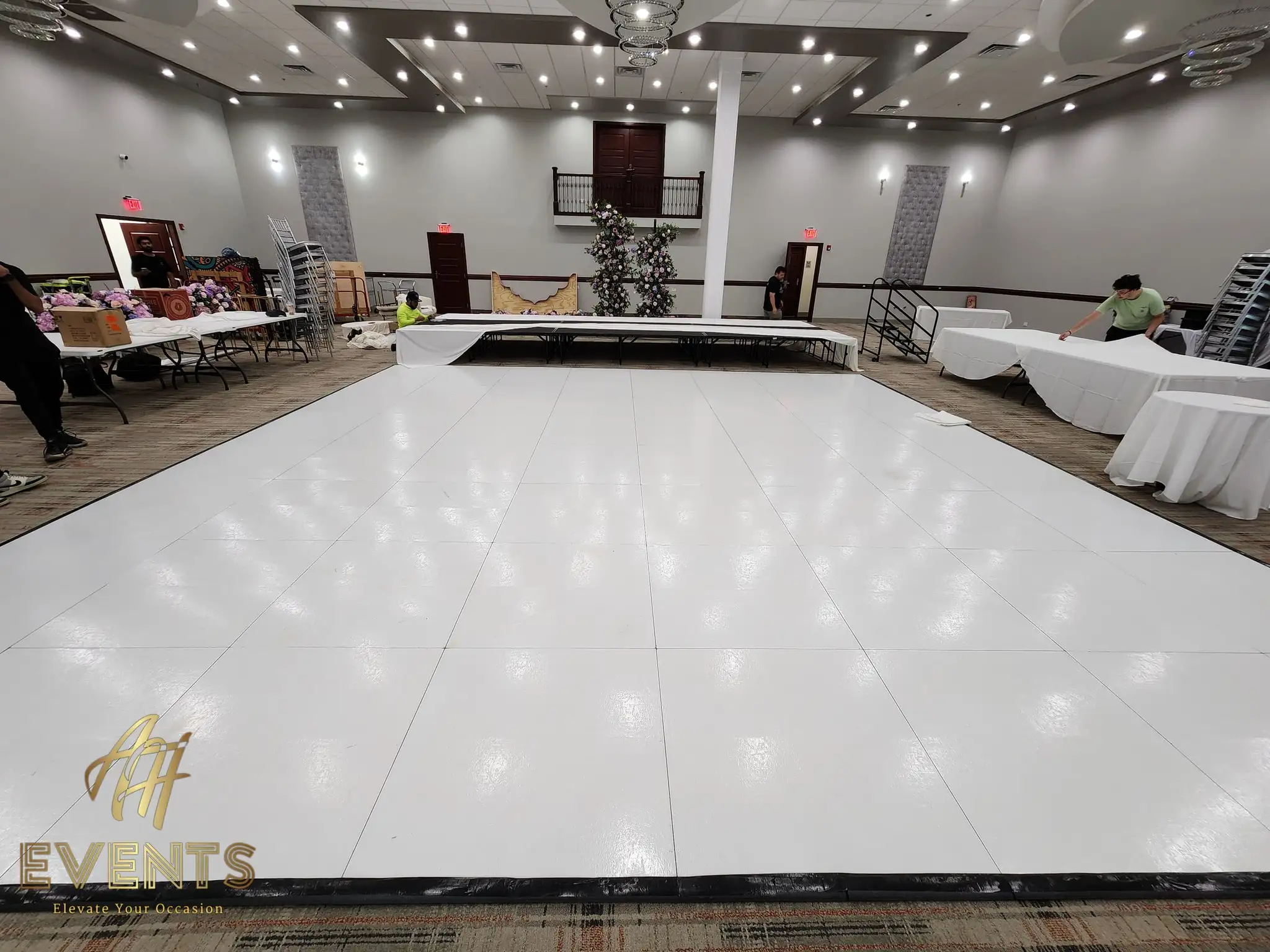 White dance floor being laid in a banquet hall during setup, with a low floral stage behind