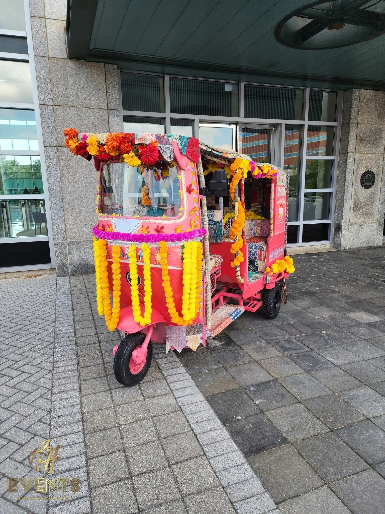 Auto rickshaw draped in yellow and orange marigold garlands outside a glass-fronted building