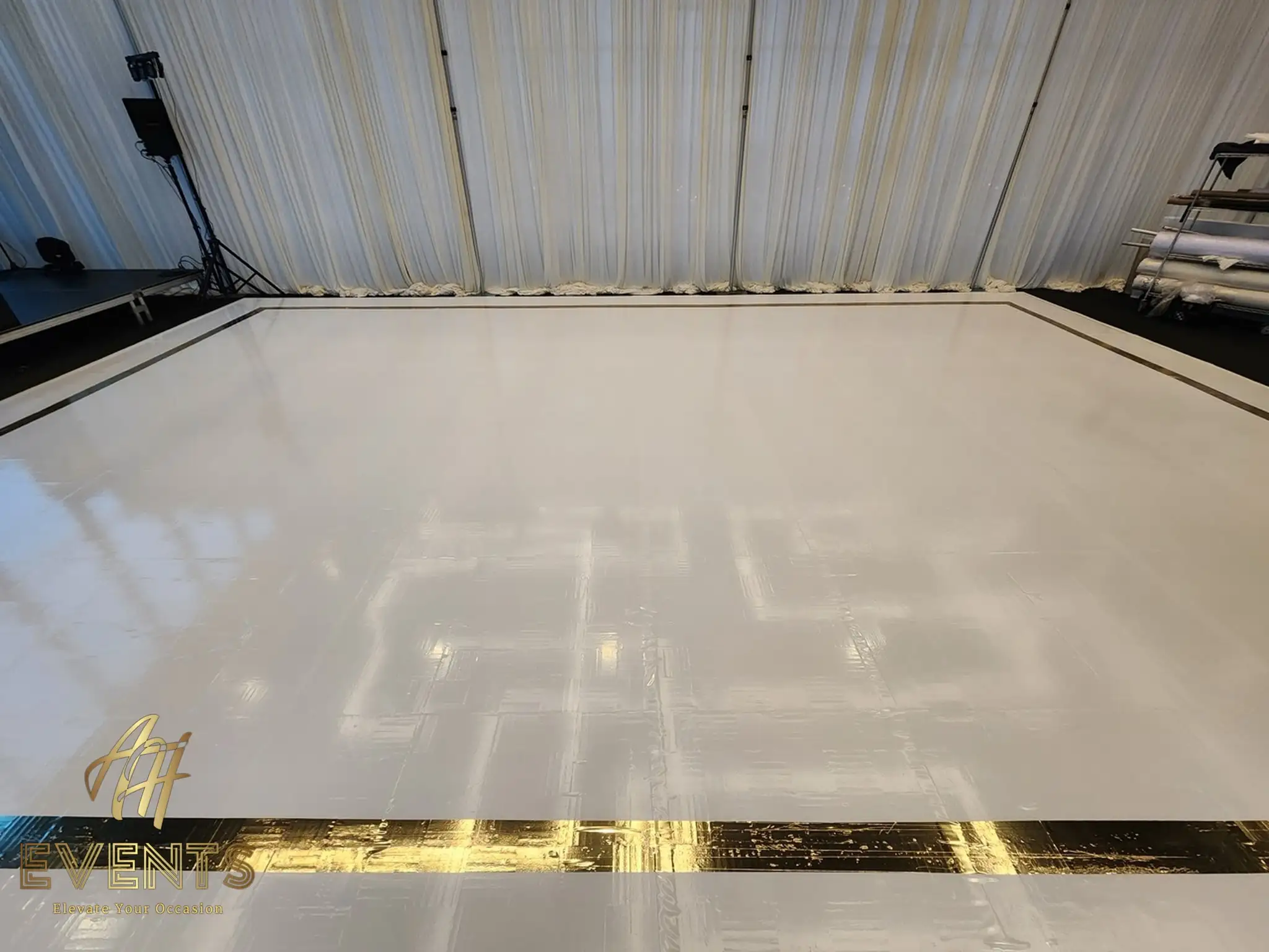 White dance floor with a gold mirror-stripe border being set up against a white pleated drape, vinyl rolls nearby
