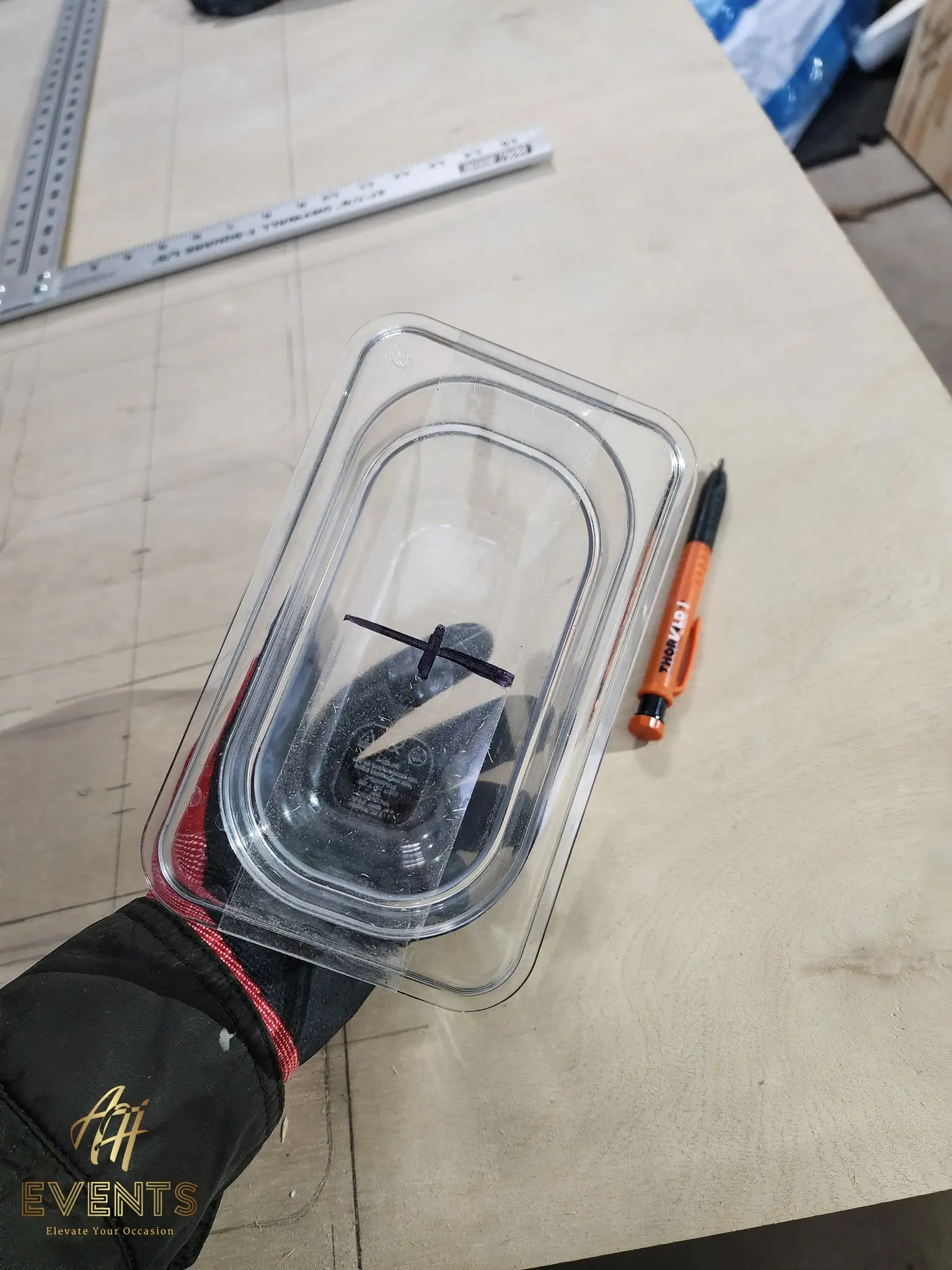 A clear plastic food-pan container used as a template on a plywood worktable beside a carpenter's square and marker