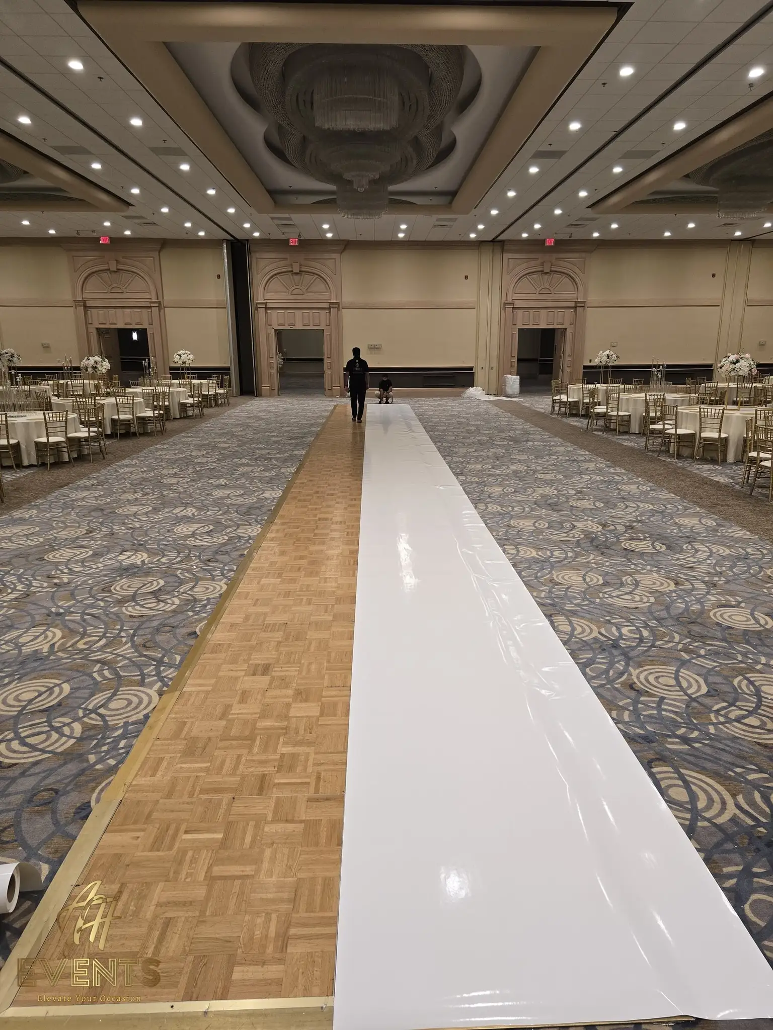 White vinyl aisle runner partway rolled out over a parquet dance floor in a ballroom during setup