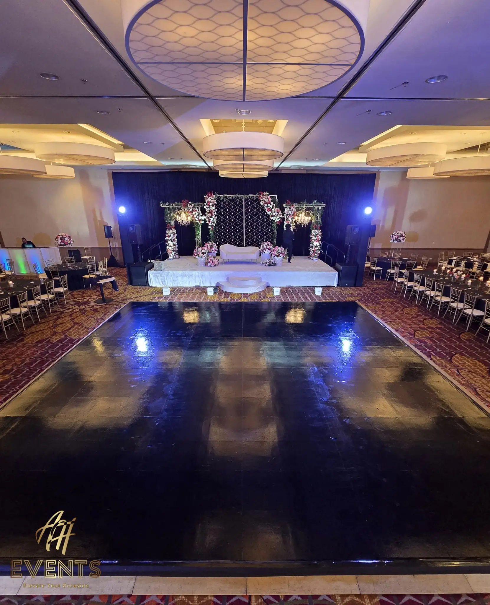 Wide ballroom view of the black dance floor leading to a white floral mandap stage with rows of chairs