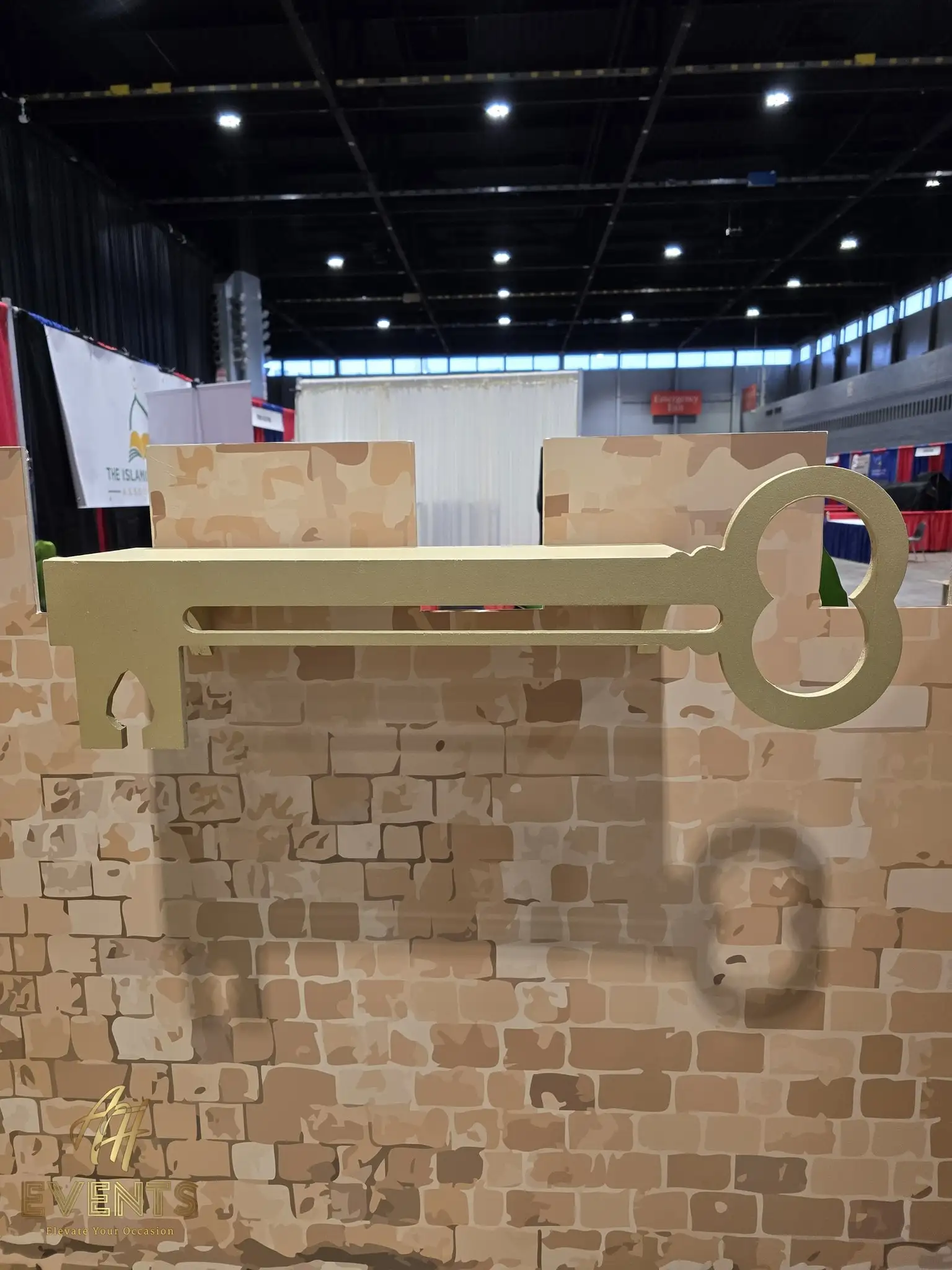 Gold key fabricated on the brick-print booth wall, the convention hall in the background