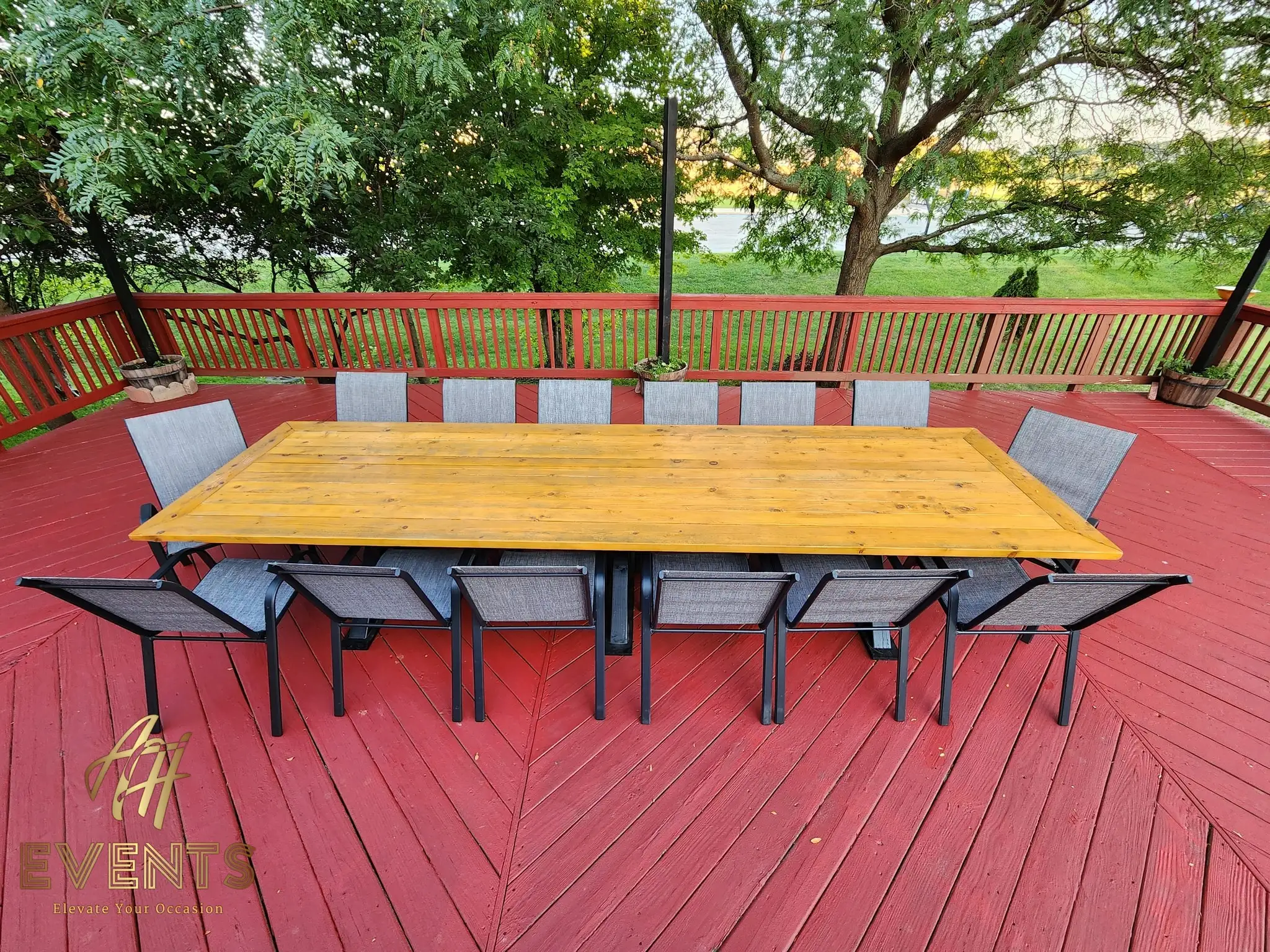 Long wooden farm table with gray chairs centered on a red deck, framed by trees