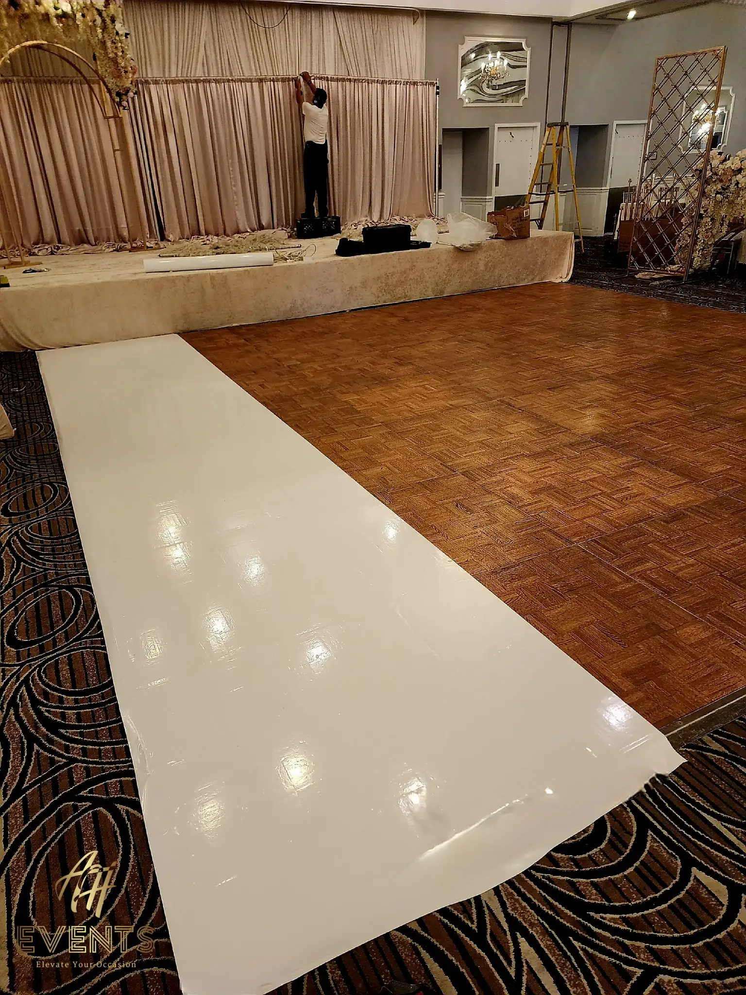 White vinyl dance floor partly unrolled over parquet during setup, an installer hanging drape behind