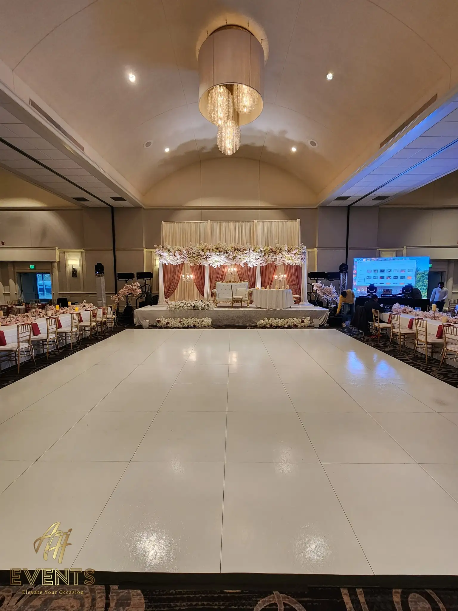 White dance floor under a drum chandelier, facing a blush stage with cherry blossoms and a tufted ivory sofa