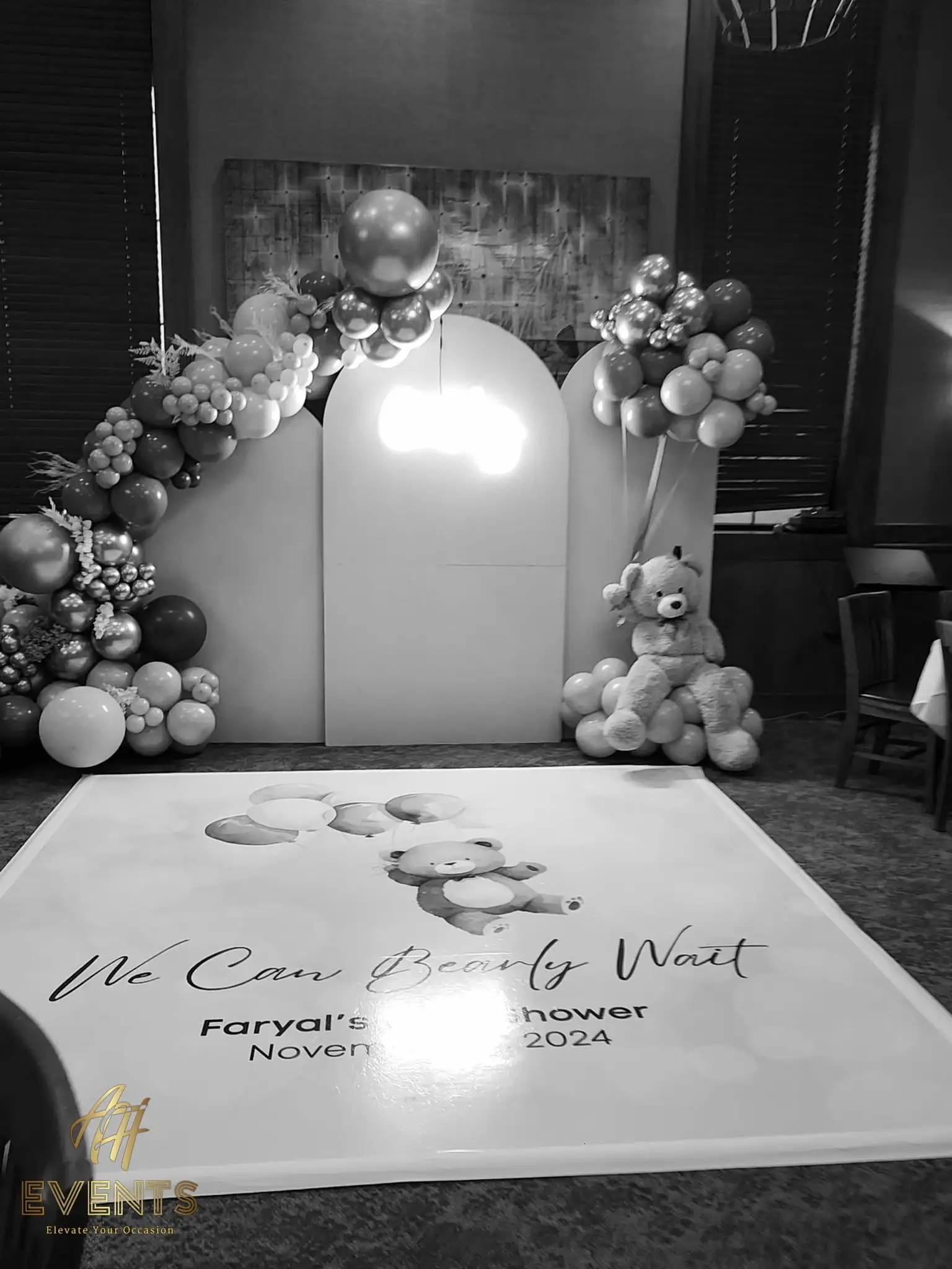 Black-and-white view of the bear-themed baby shower backdrop and printed floor with balloon garlands and a teddy bear