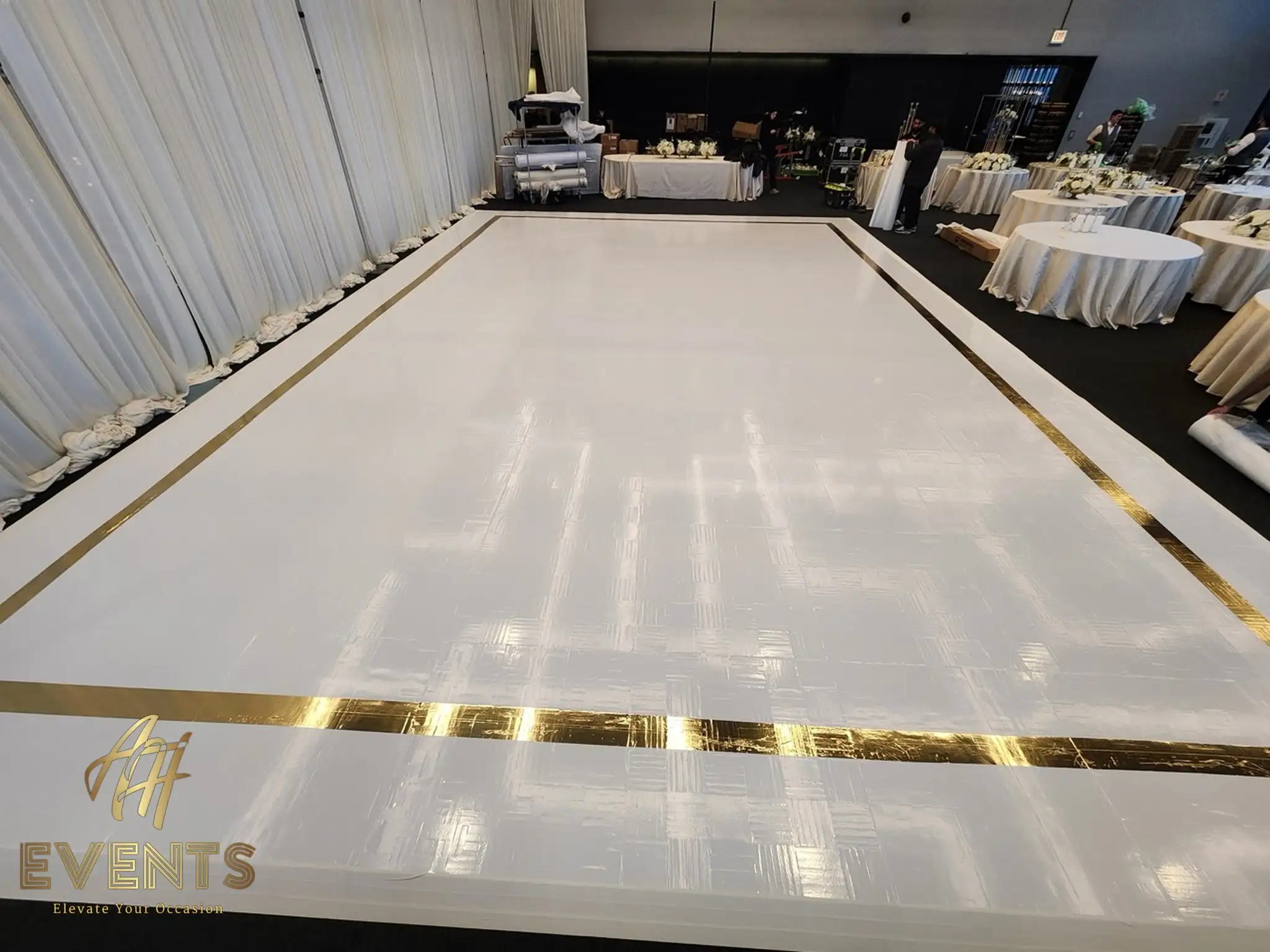 White dance floor with a gold mirror border during setup, round tables and white drape behind