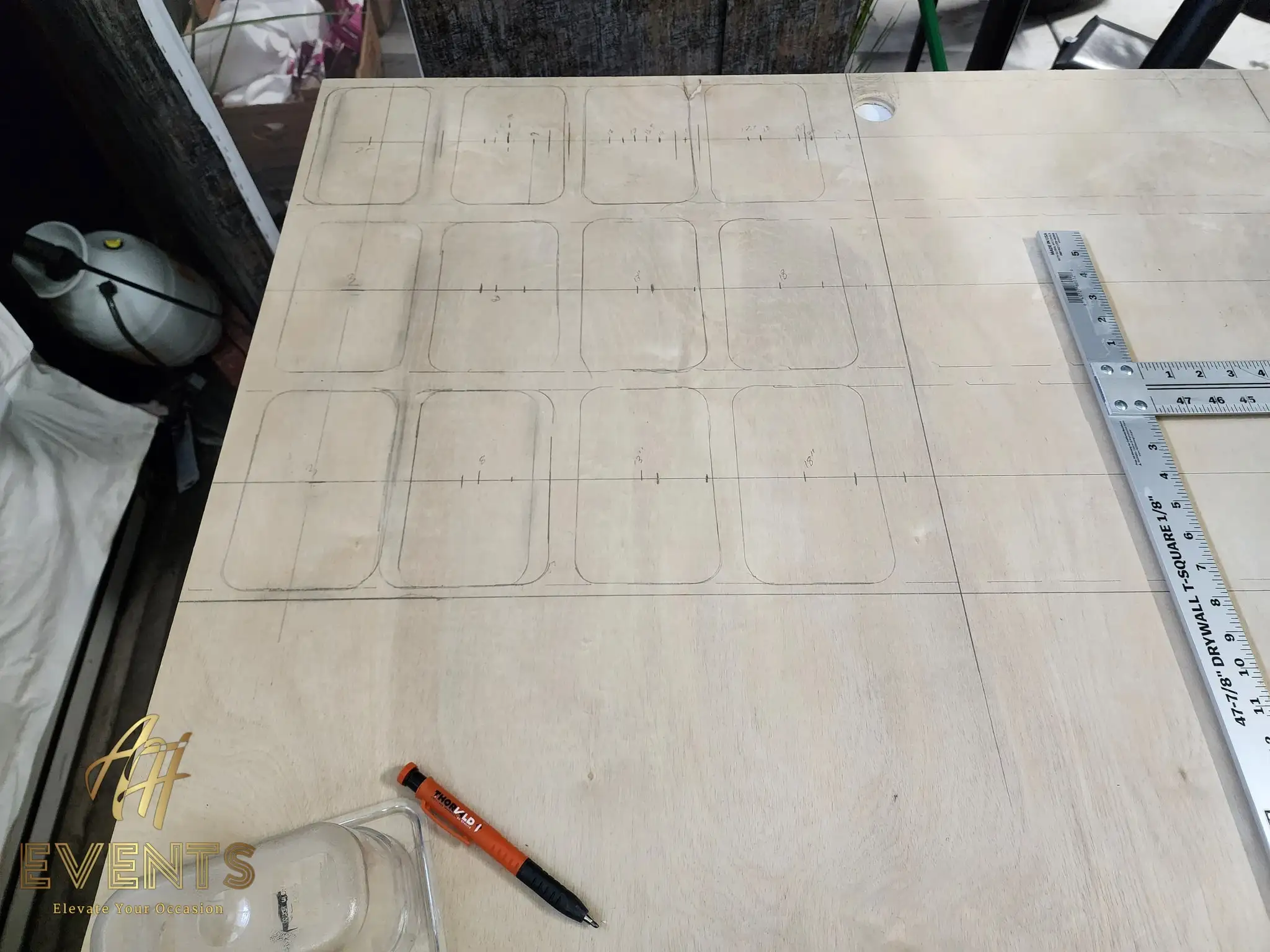 Plywood sheet marked with a pencil grid and rounded-rectangle outlines, a drywall T-square and marker laid on top