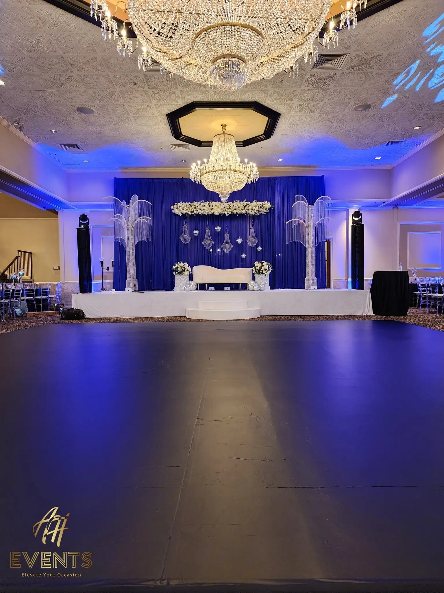 Black dance floor in a blue-lit ballroom facing a navy-draped stage with crystal palm columns and a white sofa