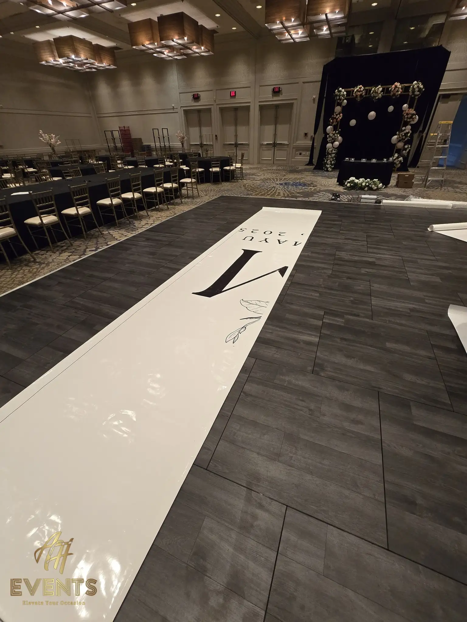A long printed monogram runner being unrolled over gray plank flooring during setup, a black floral backdrop behind