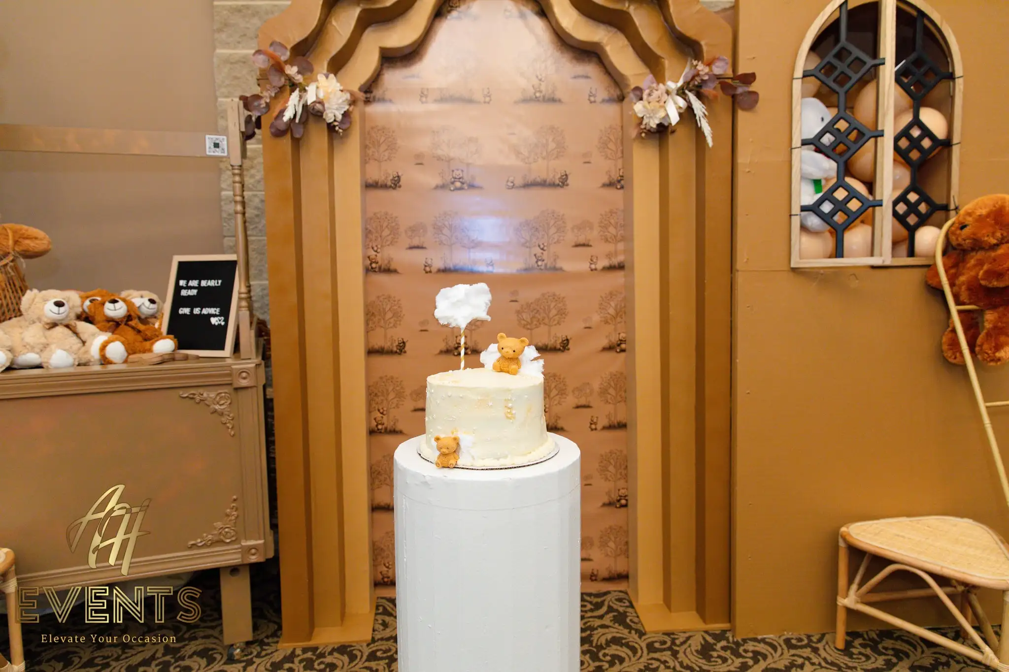 White bear-topped cake on a pedestal in front of the gold storybook house backdrop, a teddy in the arched window
