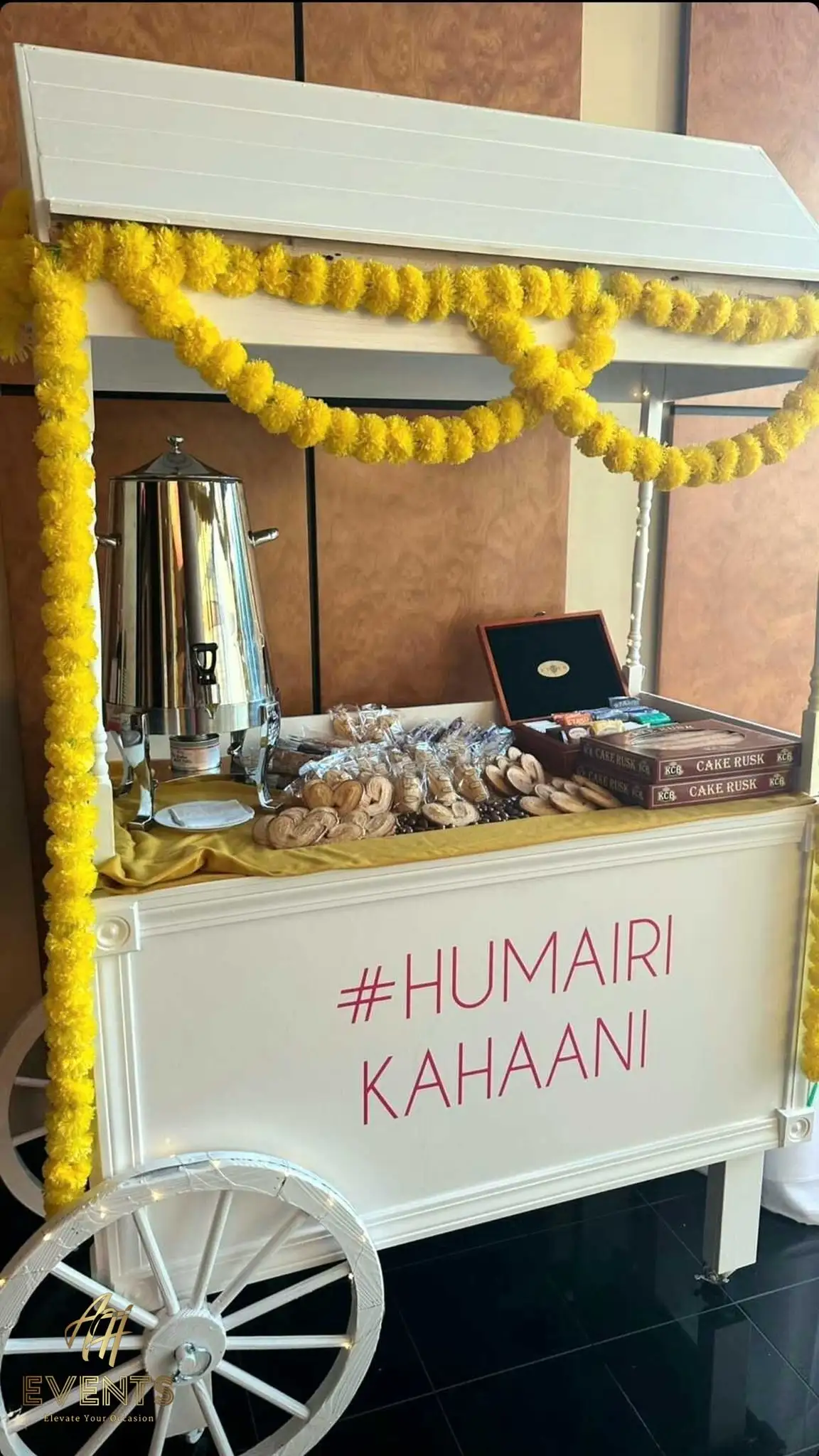 White favor cart decorated with yellow marigolds, set with a tea urn and snacks under a '#Humairi Kahaani' sign