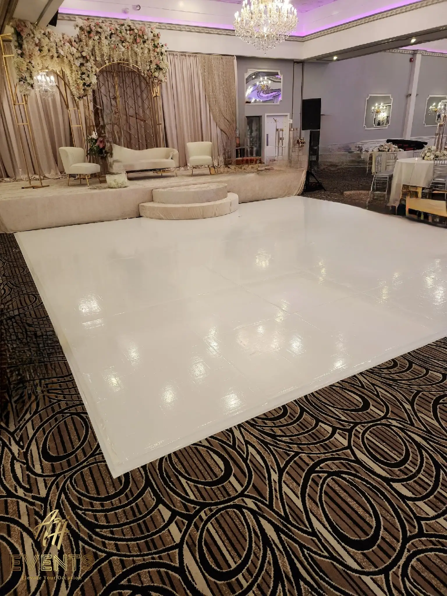 Glossy white dance floor beside a blush-and-gold stage with white sofas on a swirl-patterned carpet