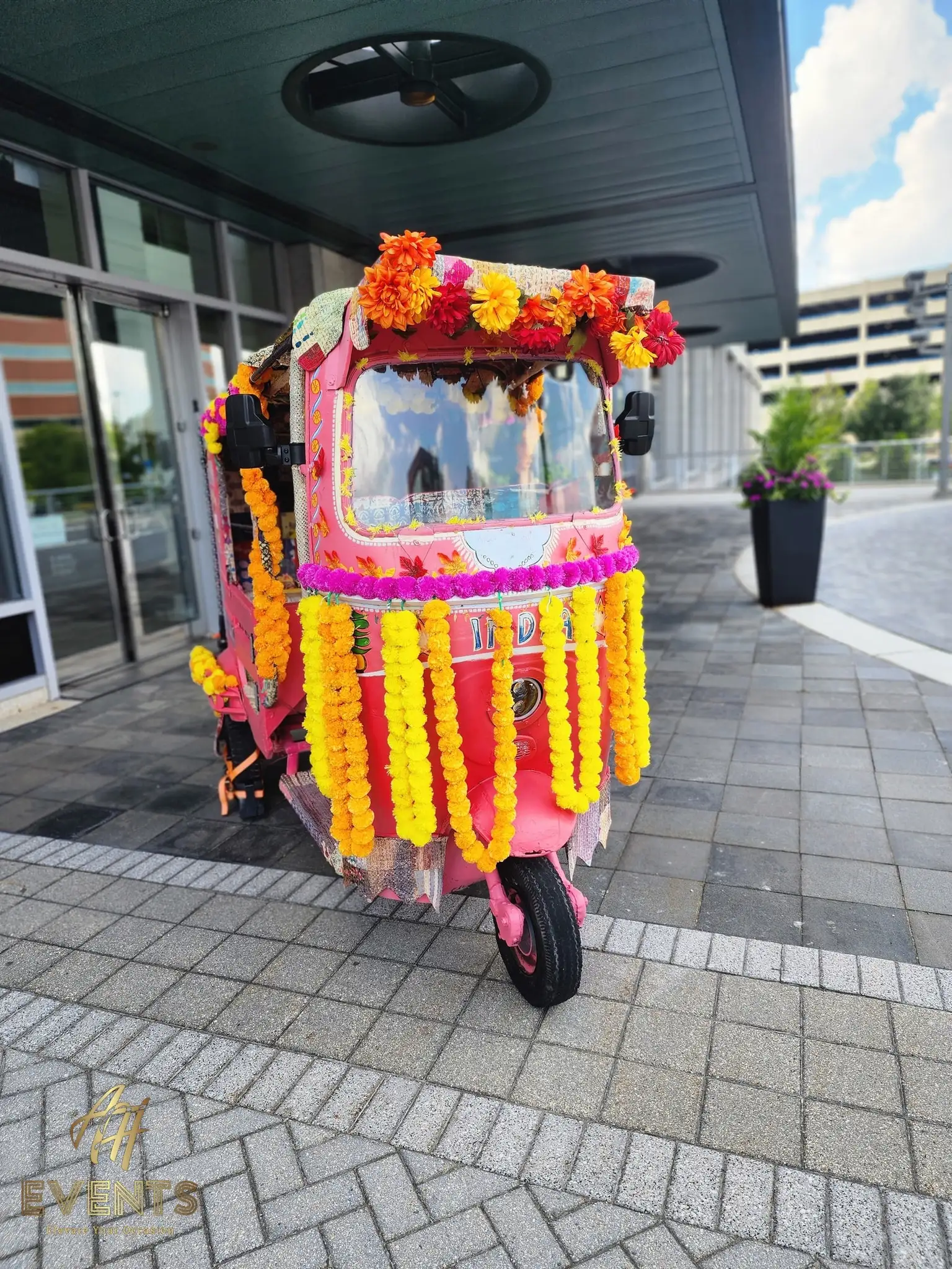 Marigold-draped auto rickshaw at a modern building entrance with a planter beside it