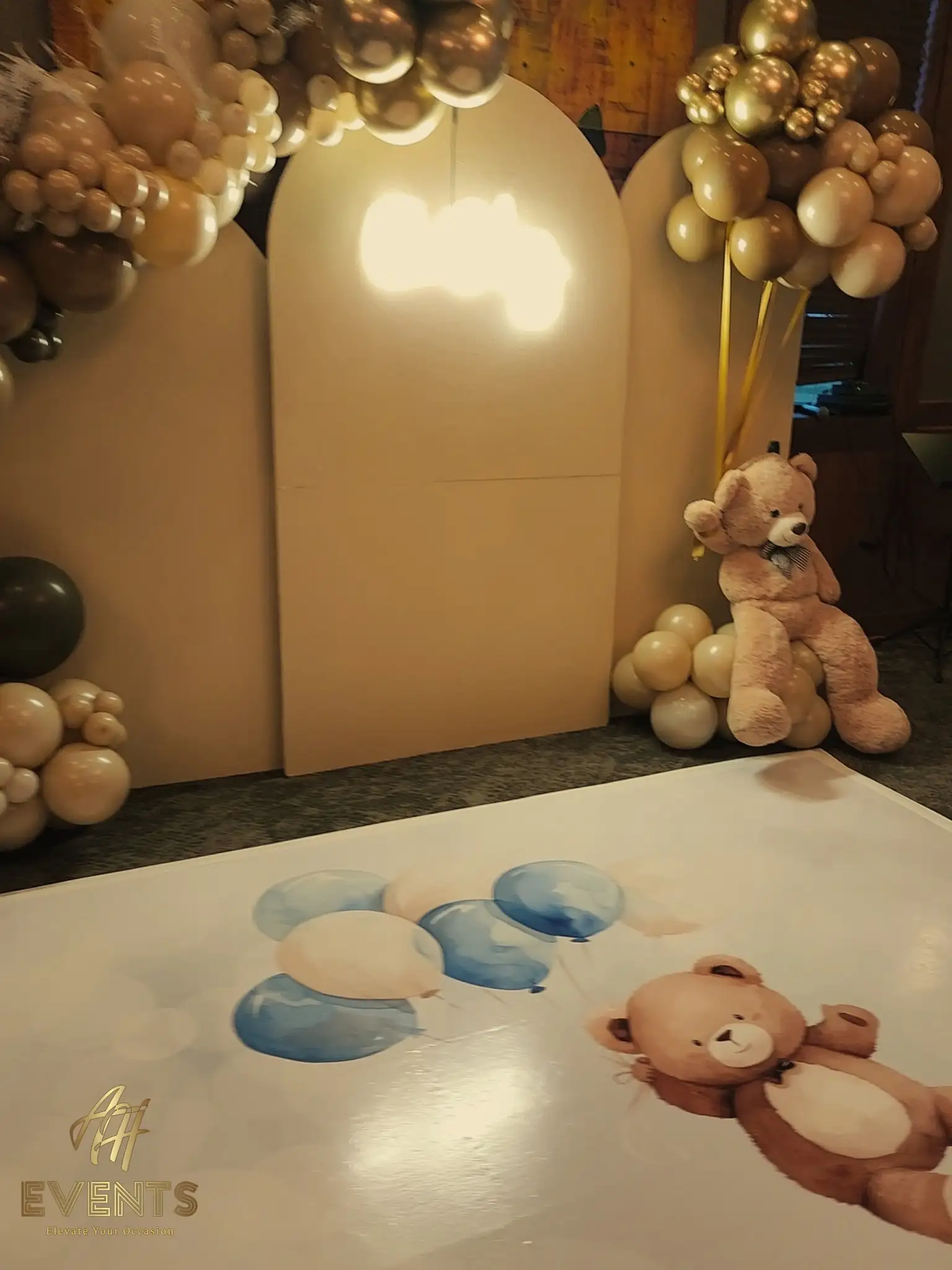 Tan arch backdrops with gold balloon clusters and a teddy bear, the printed teddy-and-balloons floor decal in front