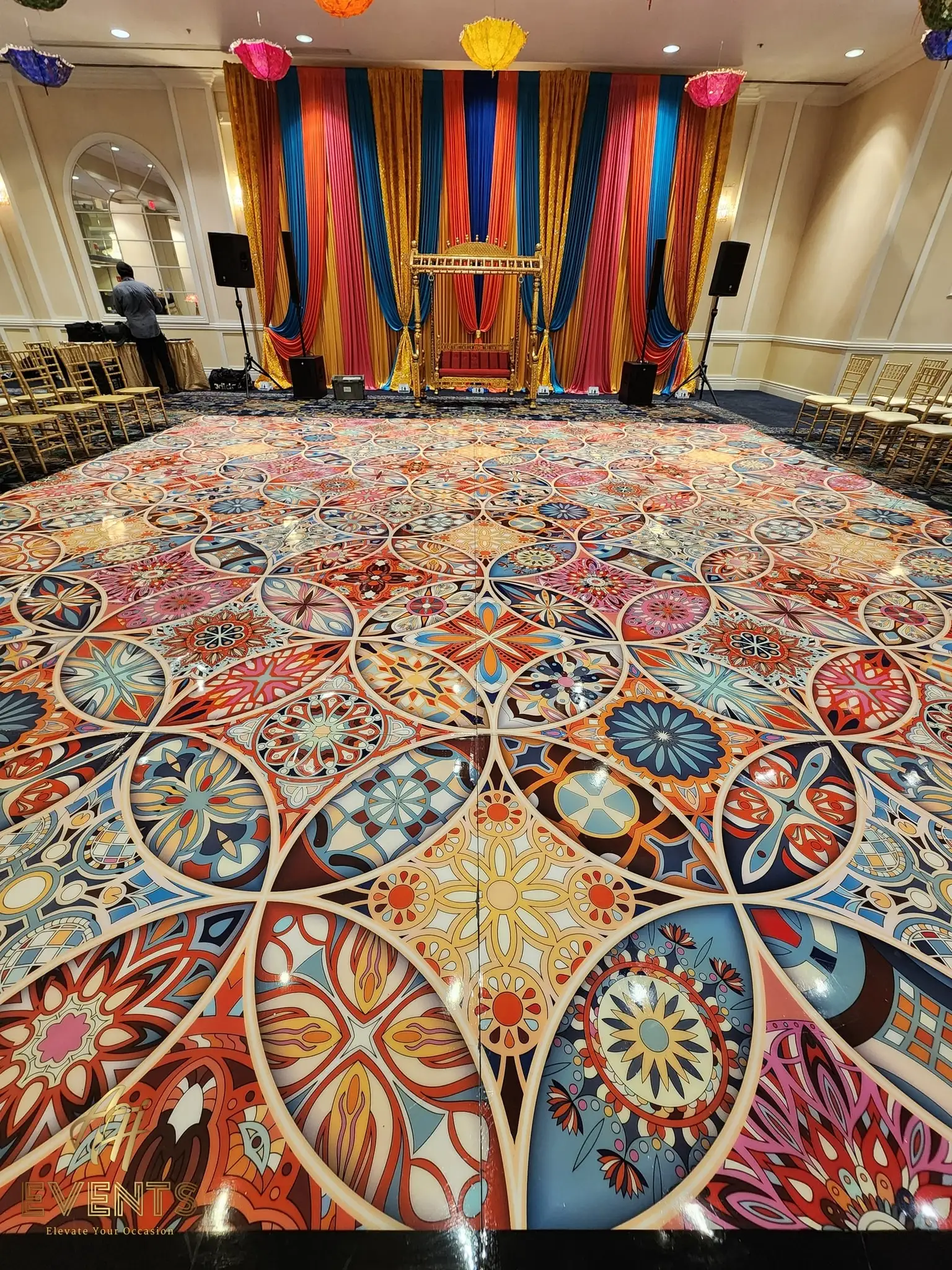 Kaleidoscope floor before a vibrant striped-drape stage in a ballroom lined with gold chairs
