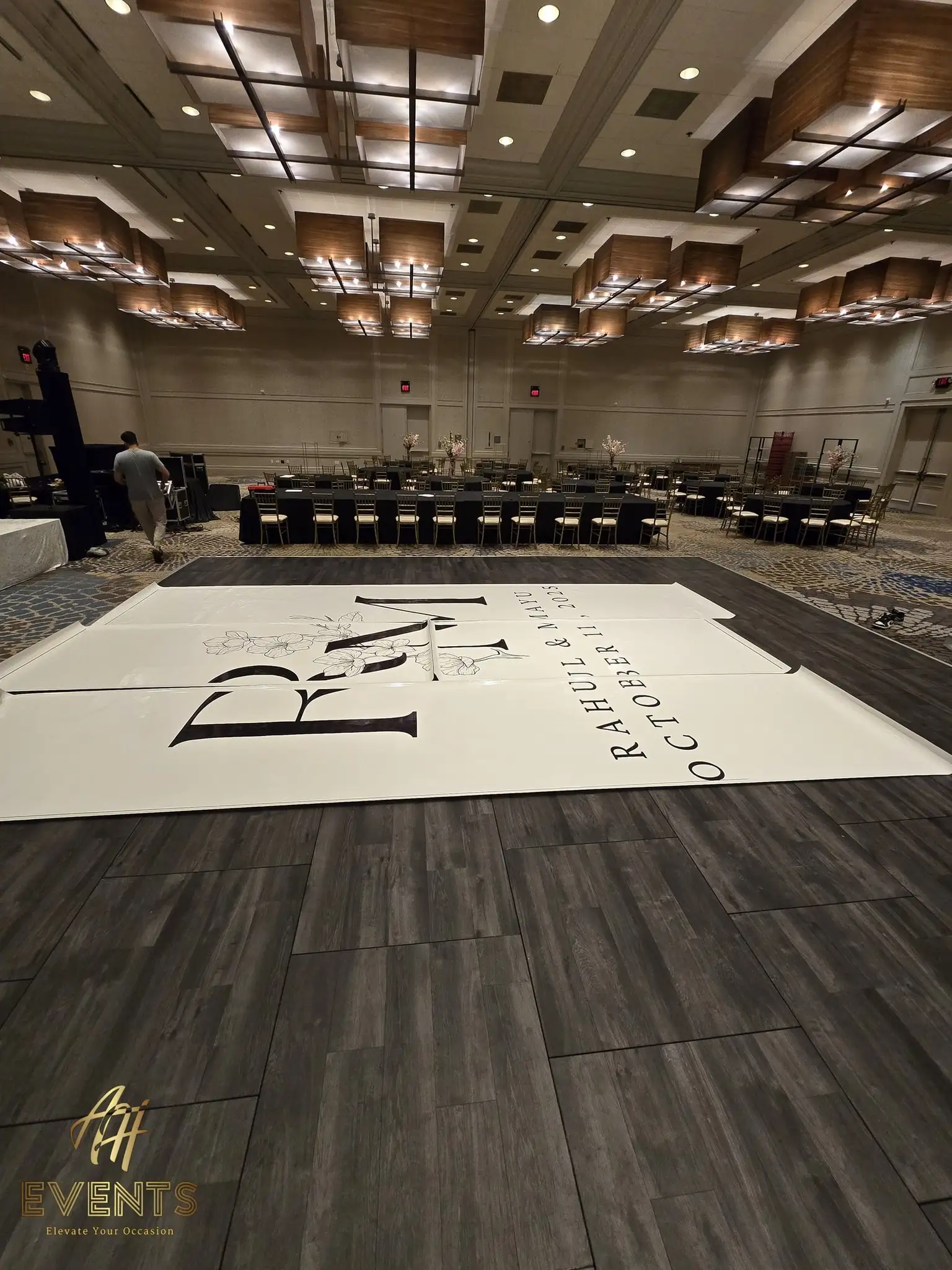The 'R M — Rahul & Mayu' monogram floor partly unrolled over gray plank flooring during setup