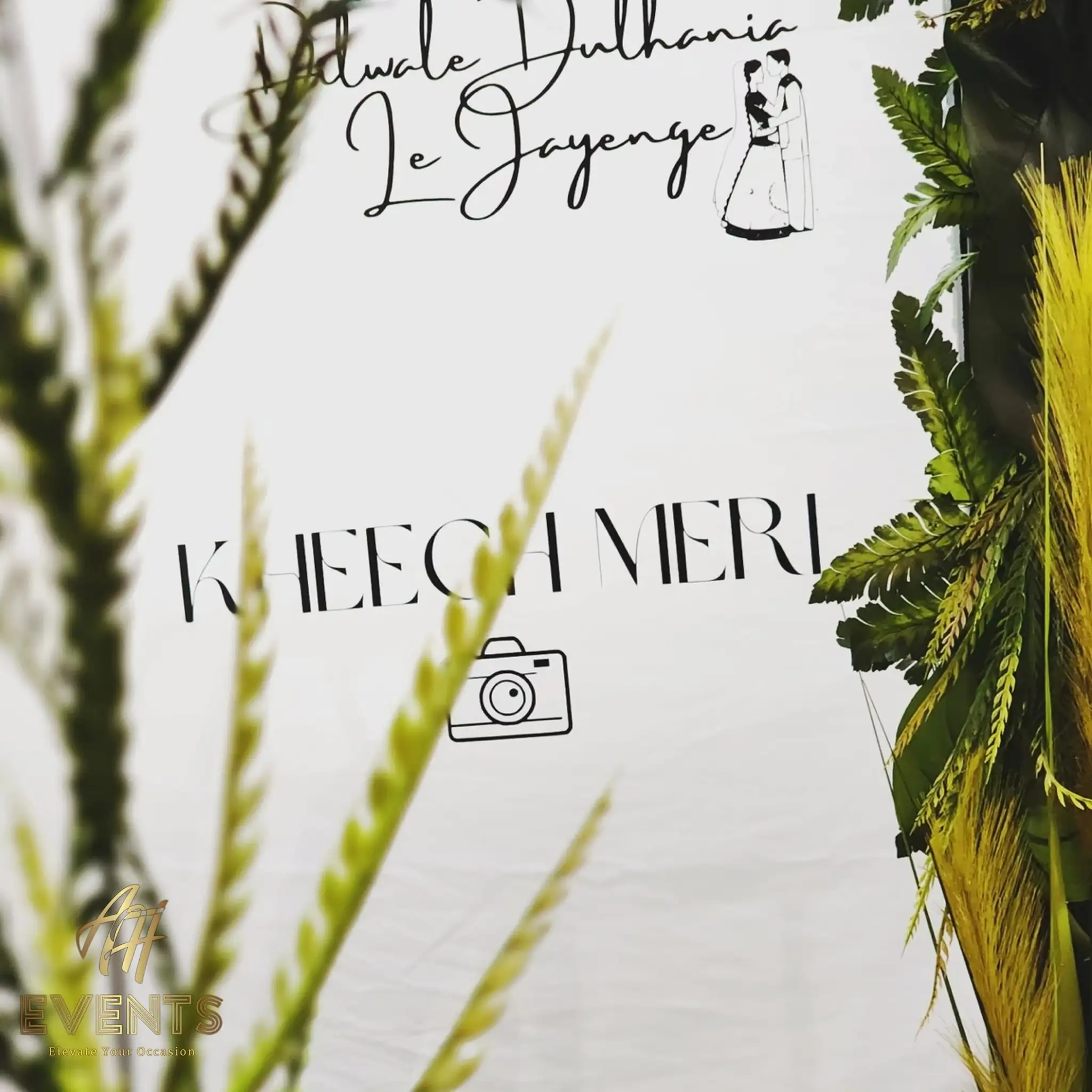 Photo-booth backdrop reading 'Kheech Meri' with a camera icon, a bride-and-groom illustration, and greenery
