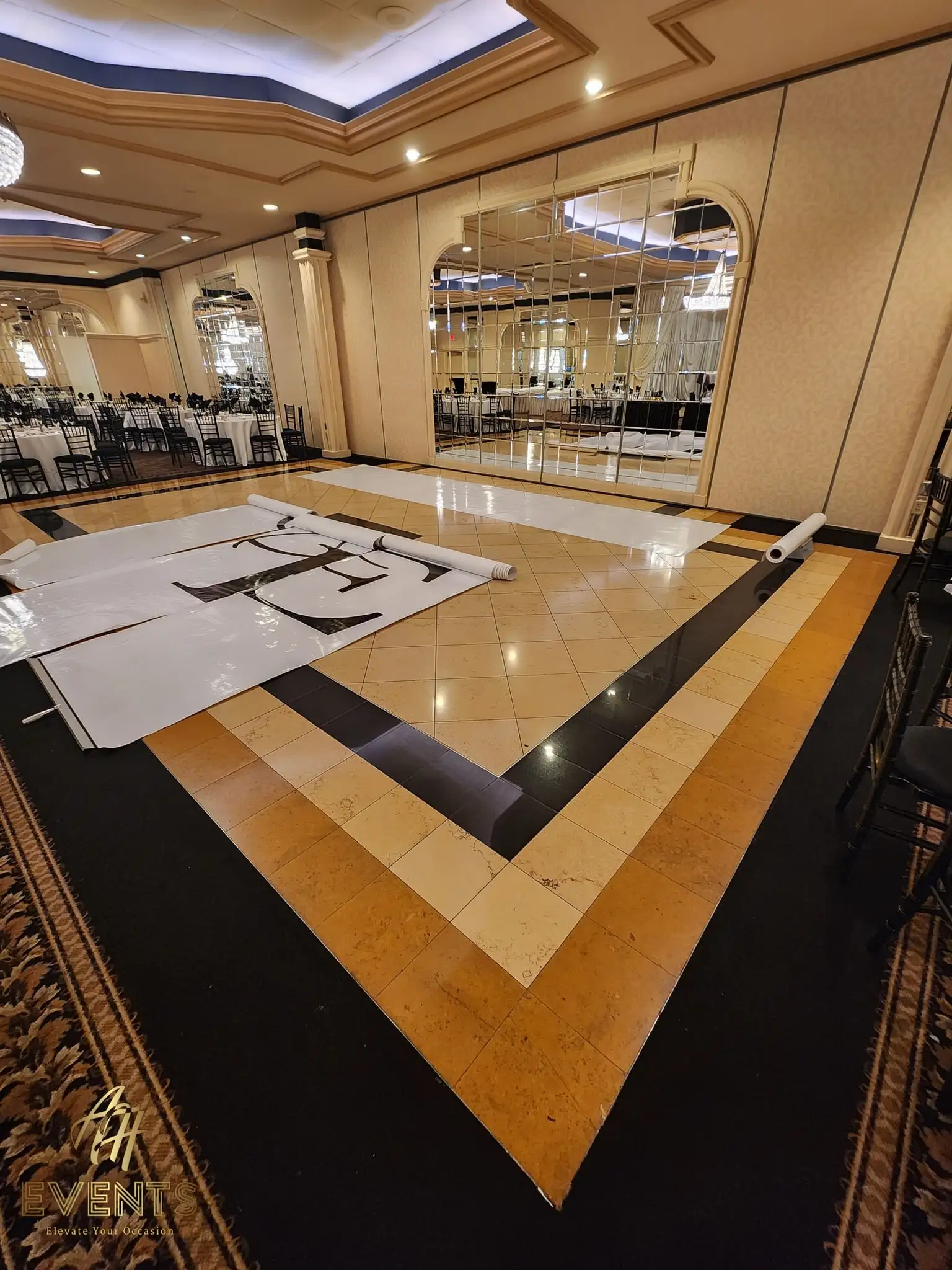 White 'A&E' monogram dance floor partly unrolled during setup, framed by tan-and-black tile and the mirrored wall