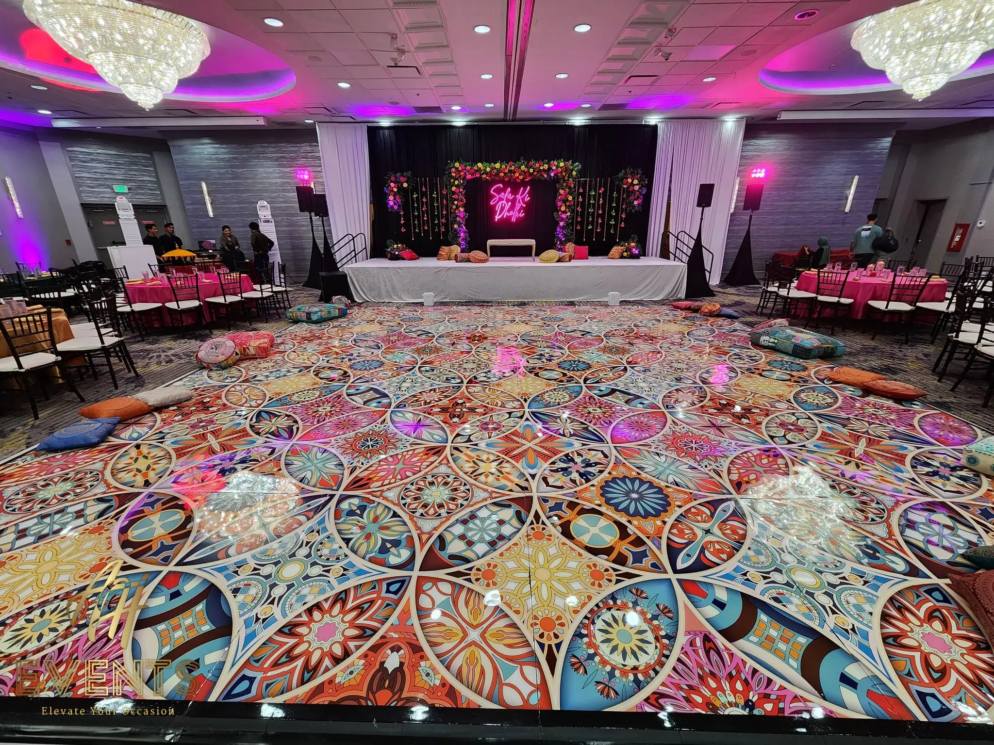 Kaleidoscope floor under purple uplighting, with floor cushions and a floral stage lit by a neon sign