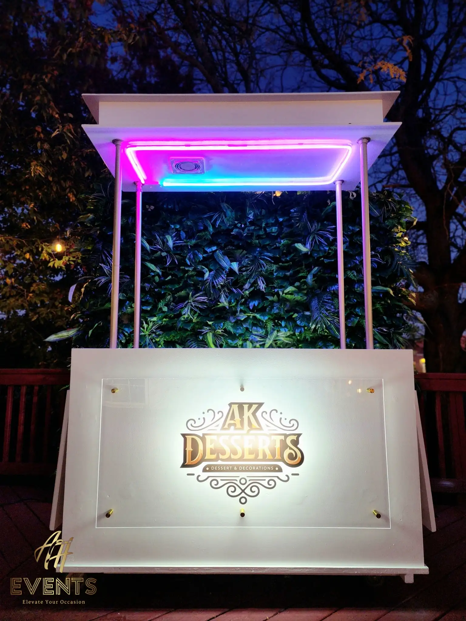 Night view of the AK Desserts cart on a deck, canopy lit pink-and-blue over a greenery wall and an illuminated logo