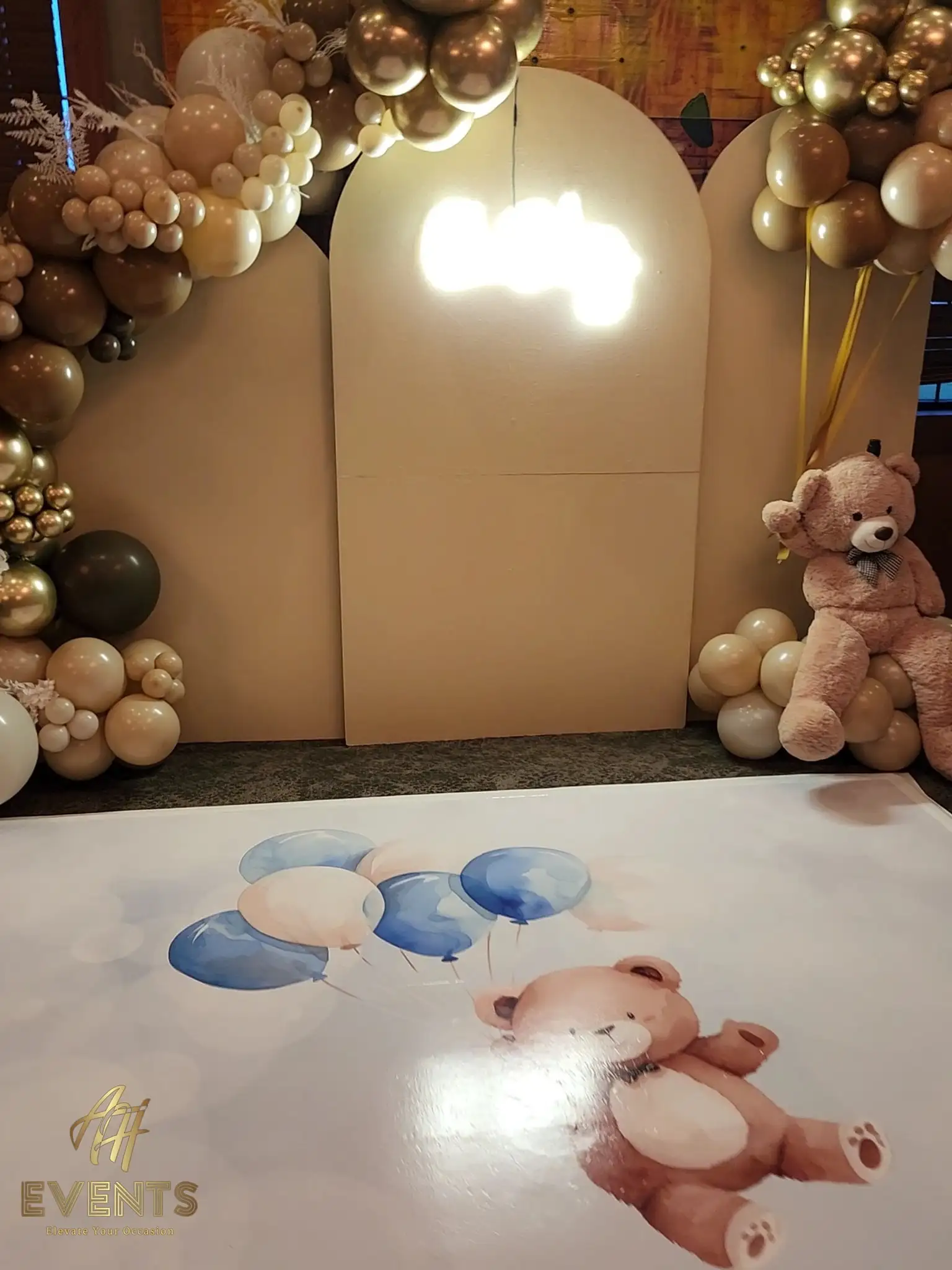 Bear-themed baby shower with beige arch backdrops, tan and gold balloon garlands, a teddy bear and a printed floor with a teddy-and-balloons illustration