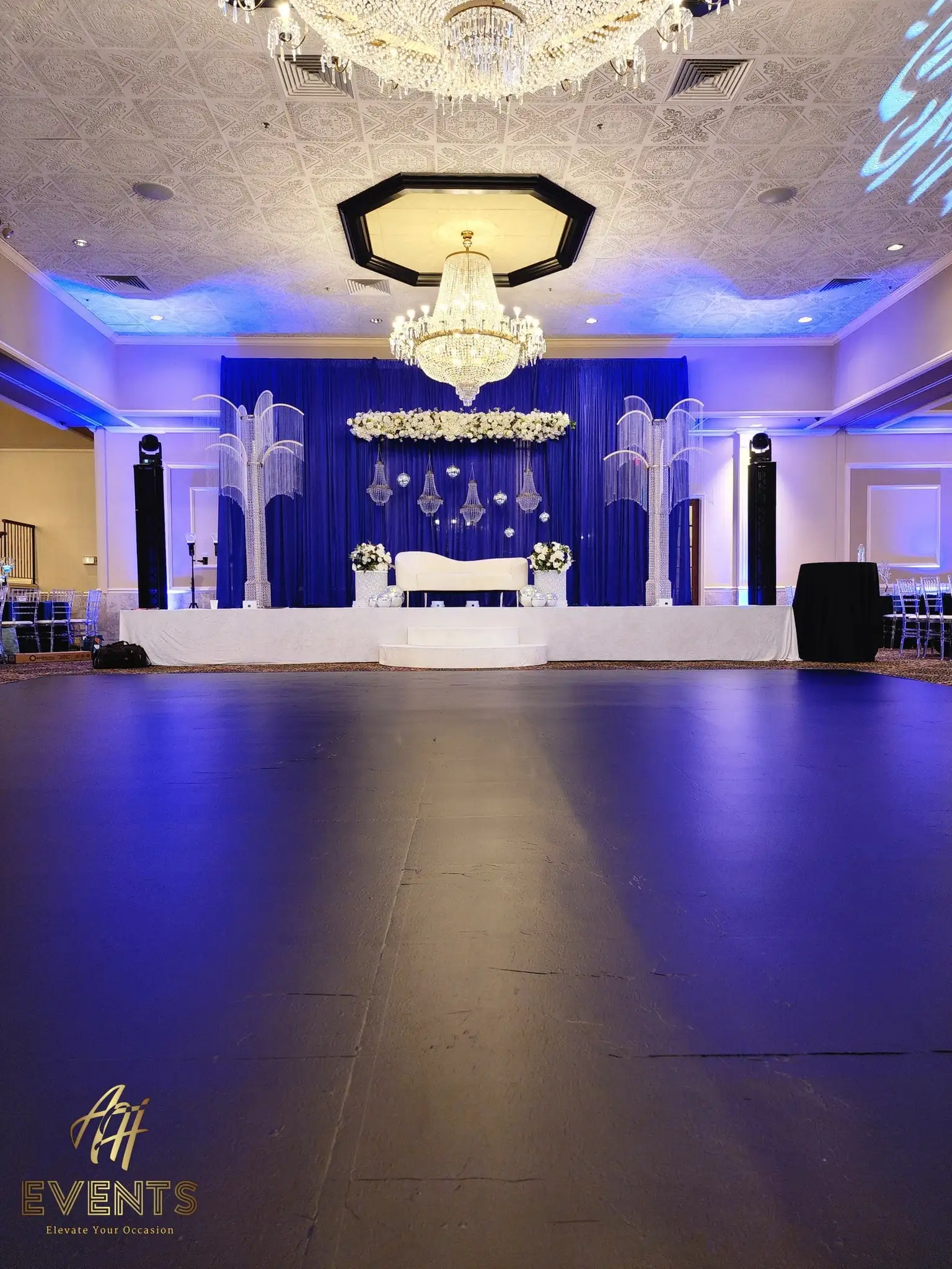 Black dance floor before a navy-draped wedding stage with a white sofa under blue uplighting