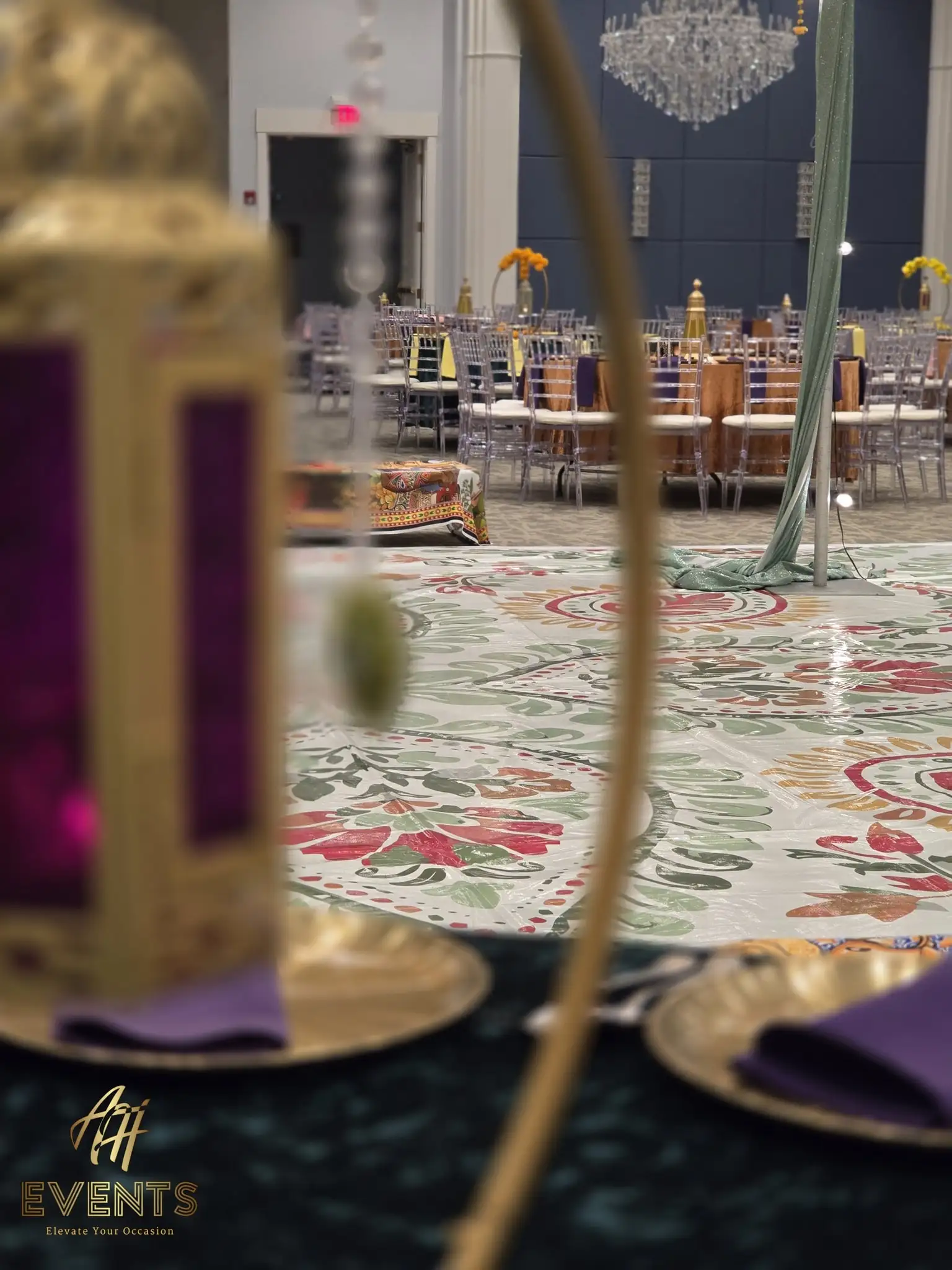 Floral flex vinyl dance floor in a banquet hall seen past a gold lantern, with a crystal chandelier and set tables