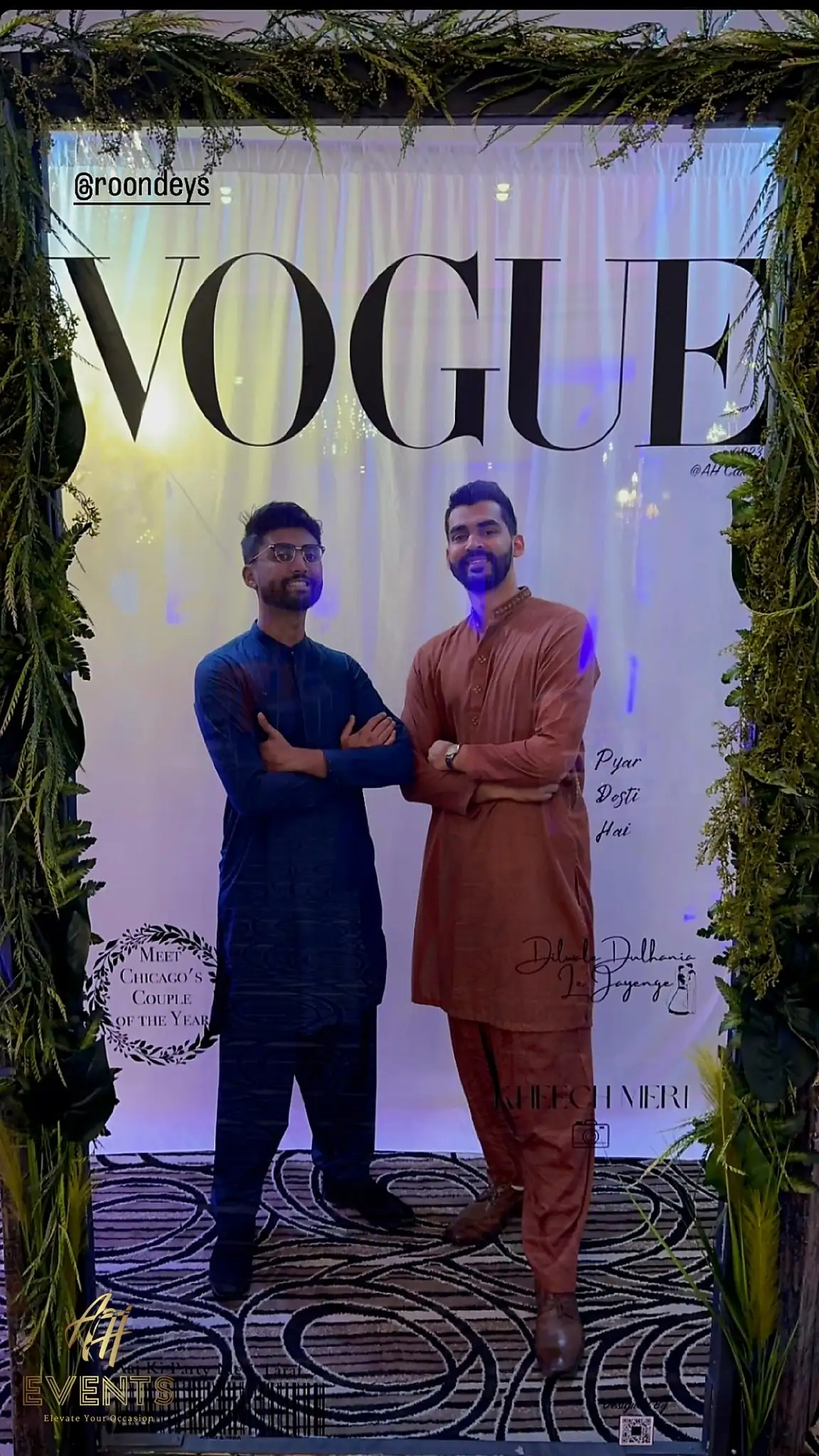 Two men posing in a VOGUE-style photo-booth frame surrounded by hanging greenery