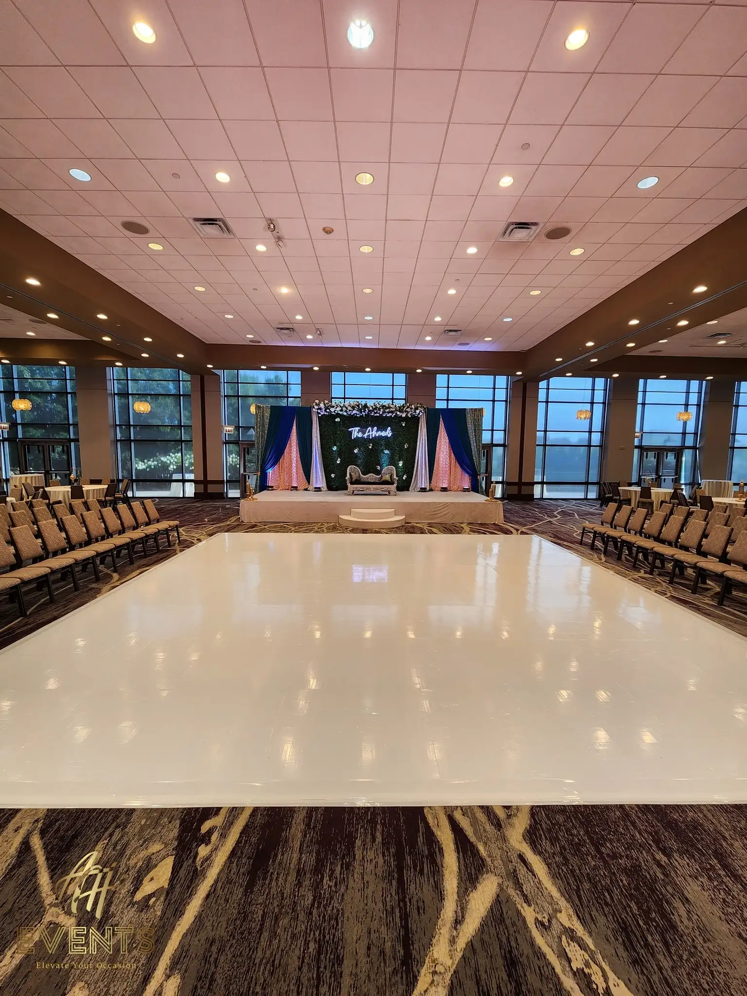 Glossy white dance floor in a window-lined ballroom facing a blue-and-pink draped stage