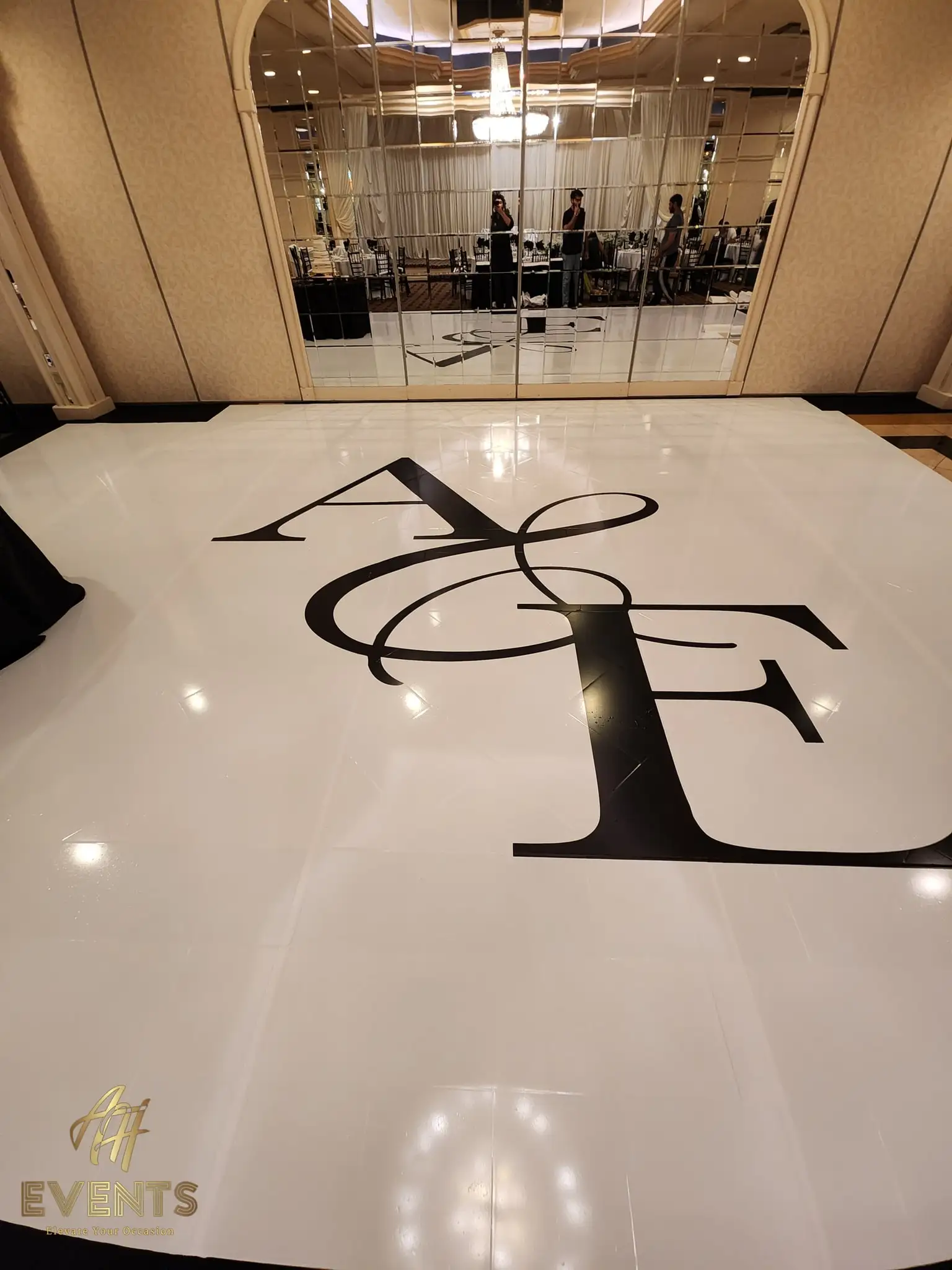 Glossy white dance floor with the large black 'A&E' serif monogram before the mirrored ballroom wall