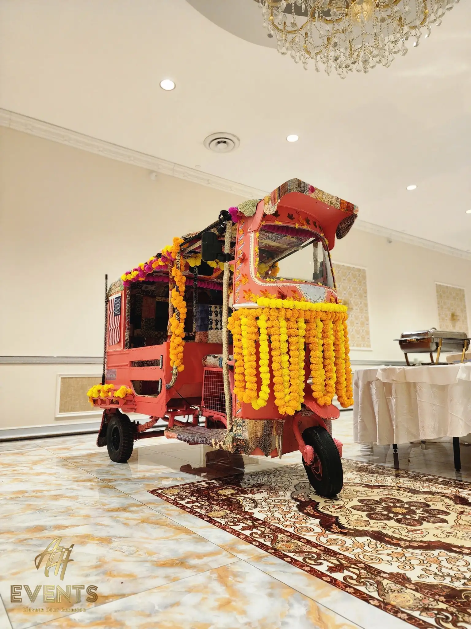 Marigold-decorated auto rickshaw in a banquet hall with a chandelier and buffet table behind