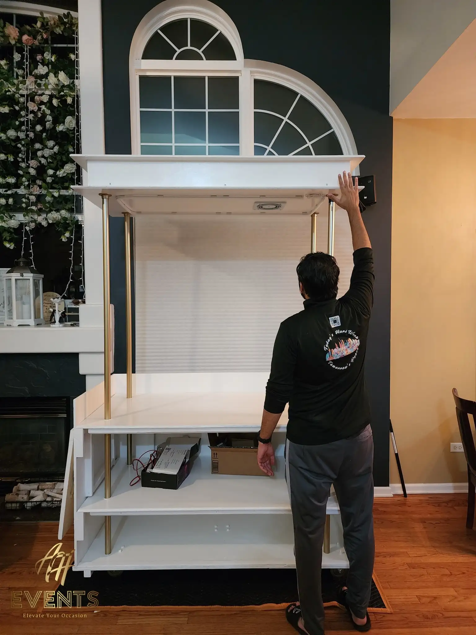 Builder assembling the white AK Desserts cart indoors, fitting the canopy onto brass poles beside a fireplace