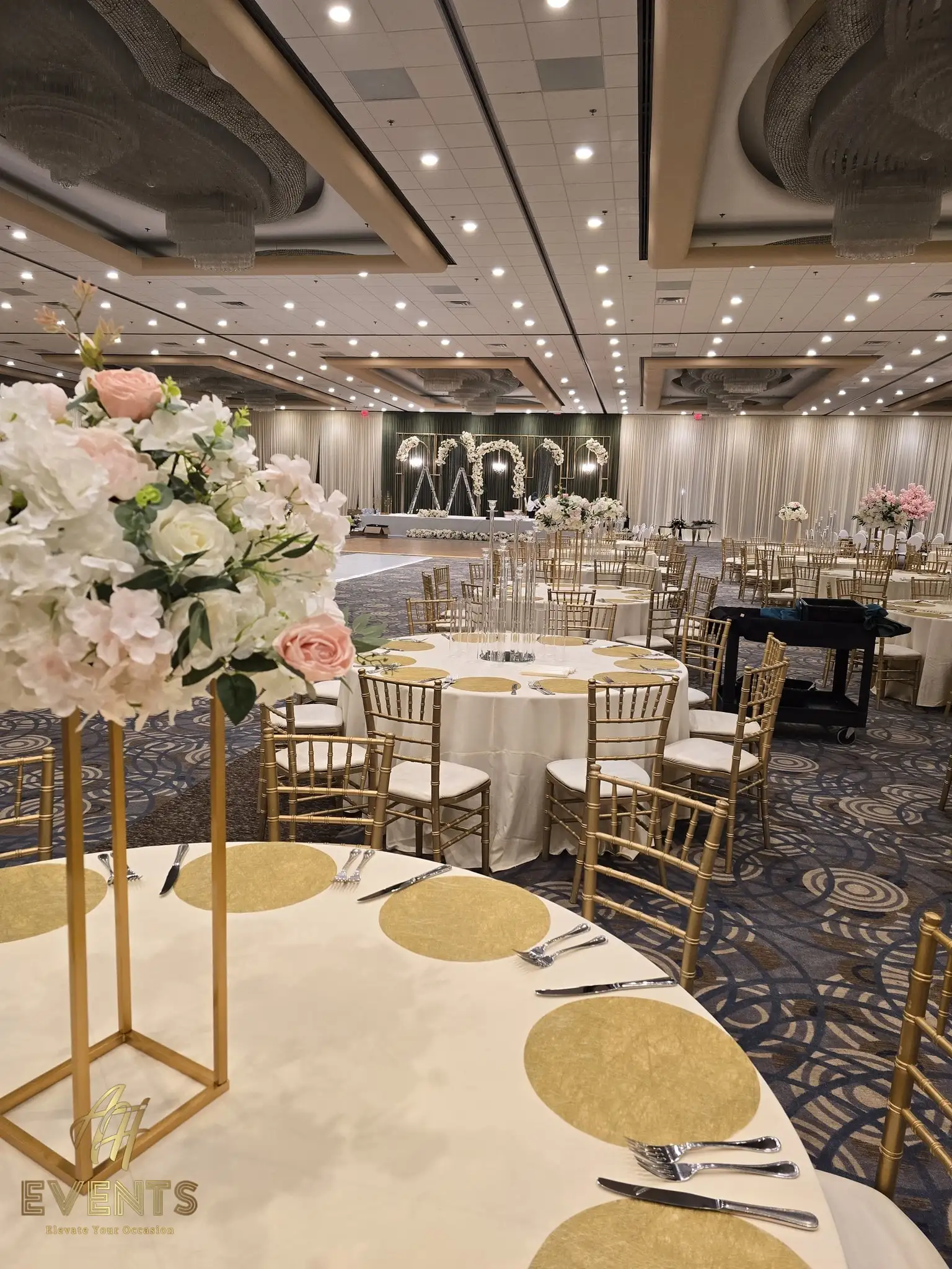 Reception table with a blush floral centerpiece and gold chargers, the white vinyl aisle and floral arch stage behind