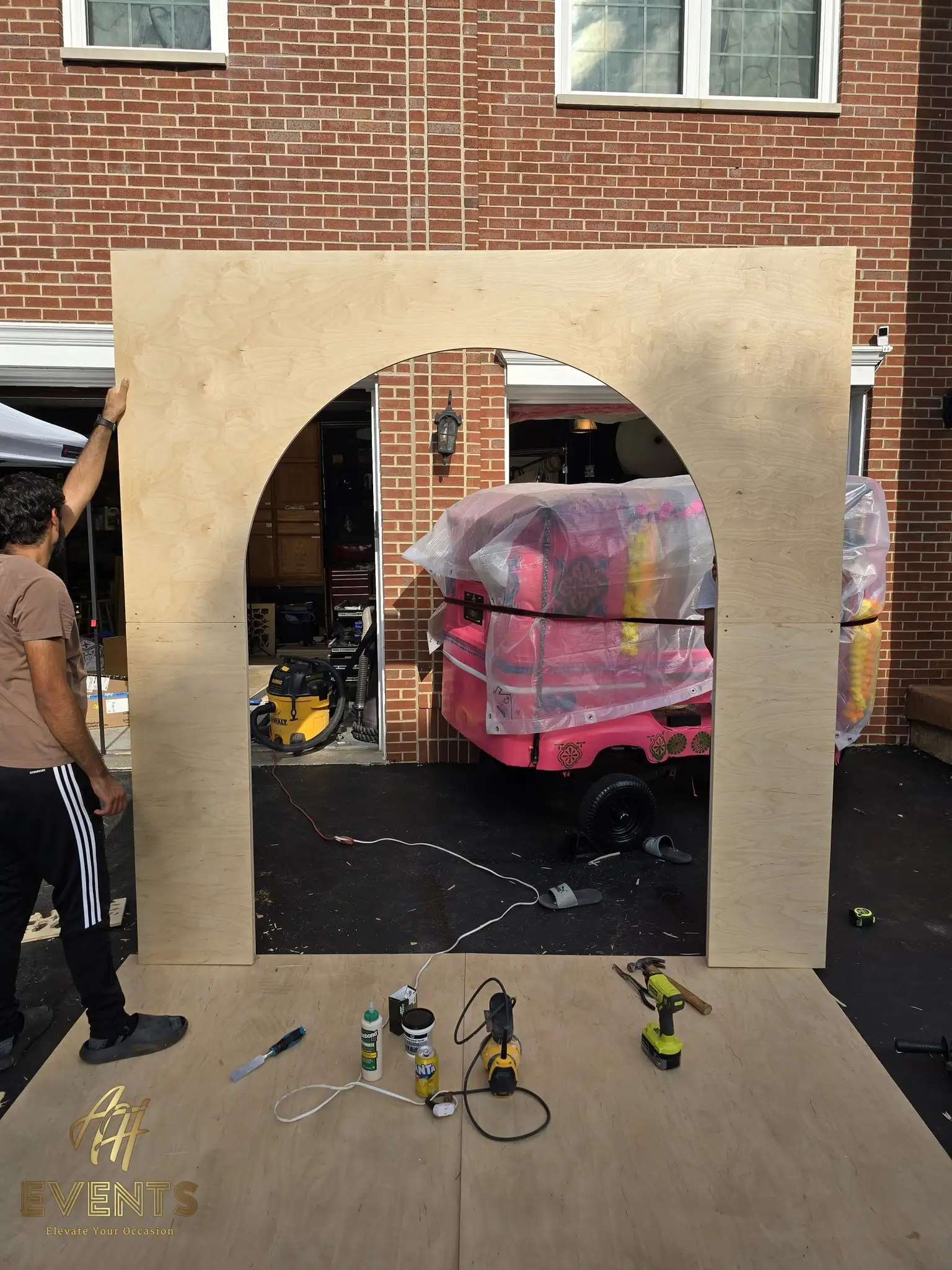 Raw plywood round-arch backdrop frame propped in a driveway, the pink auto rickshaw visible through its opening and build tools on the ground