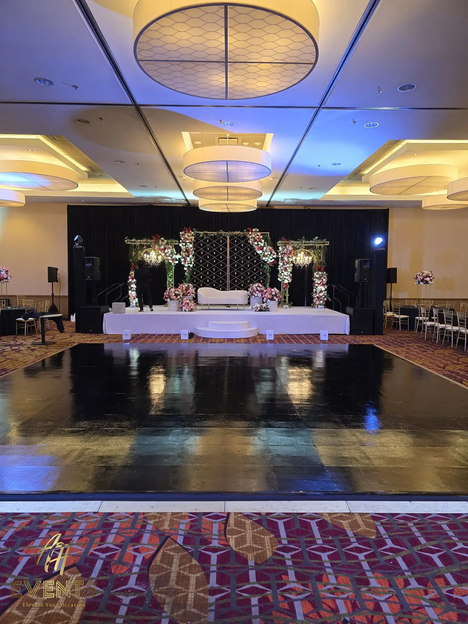 Black dance floor and white floral mandap stage framed by a patterned carpet border