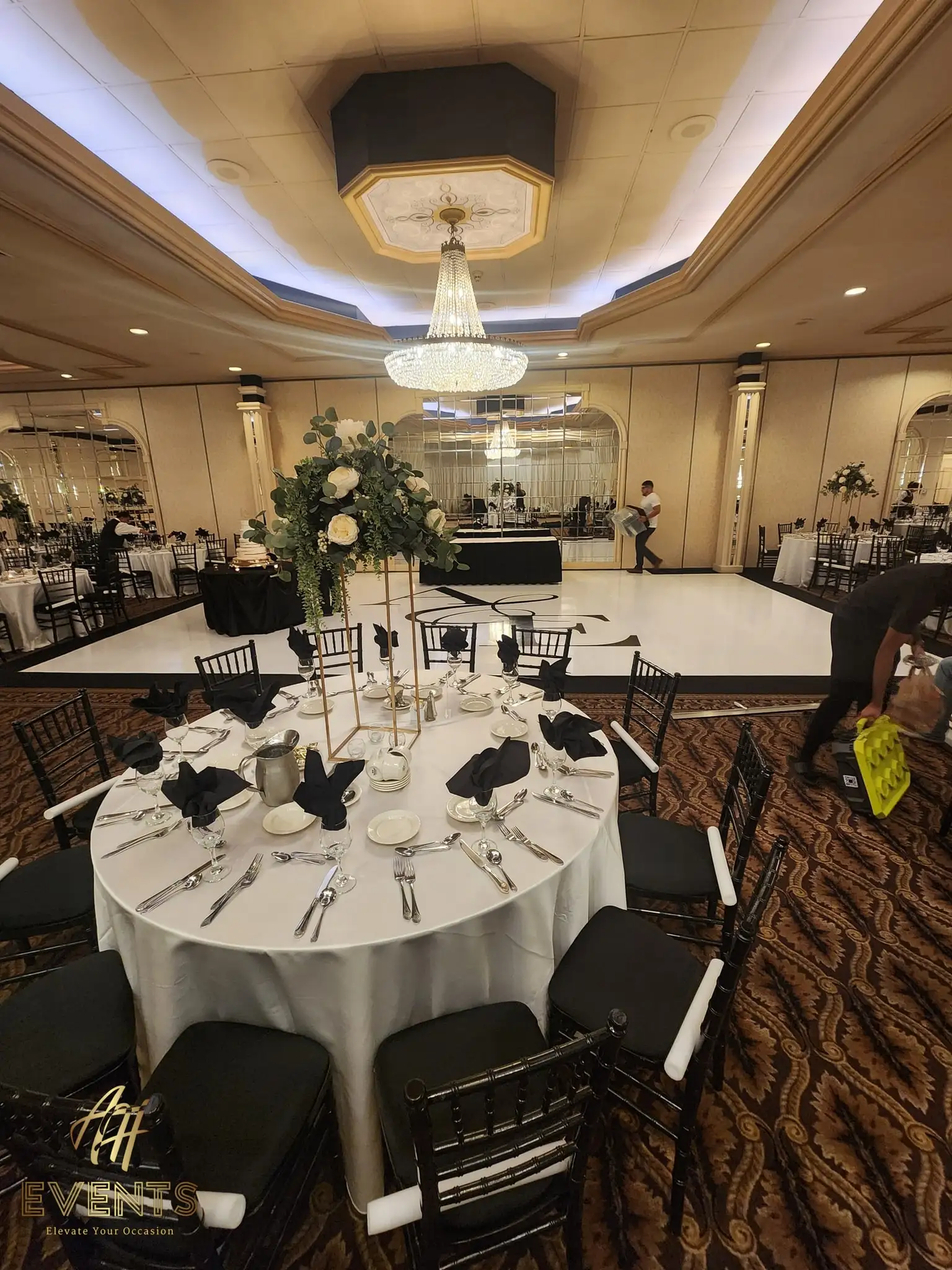Round table with a tall floral centerpiece and black chairs beside the white monogram dance floor