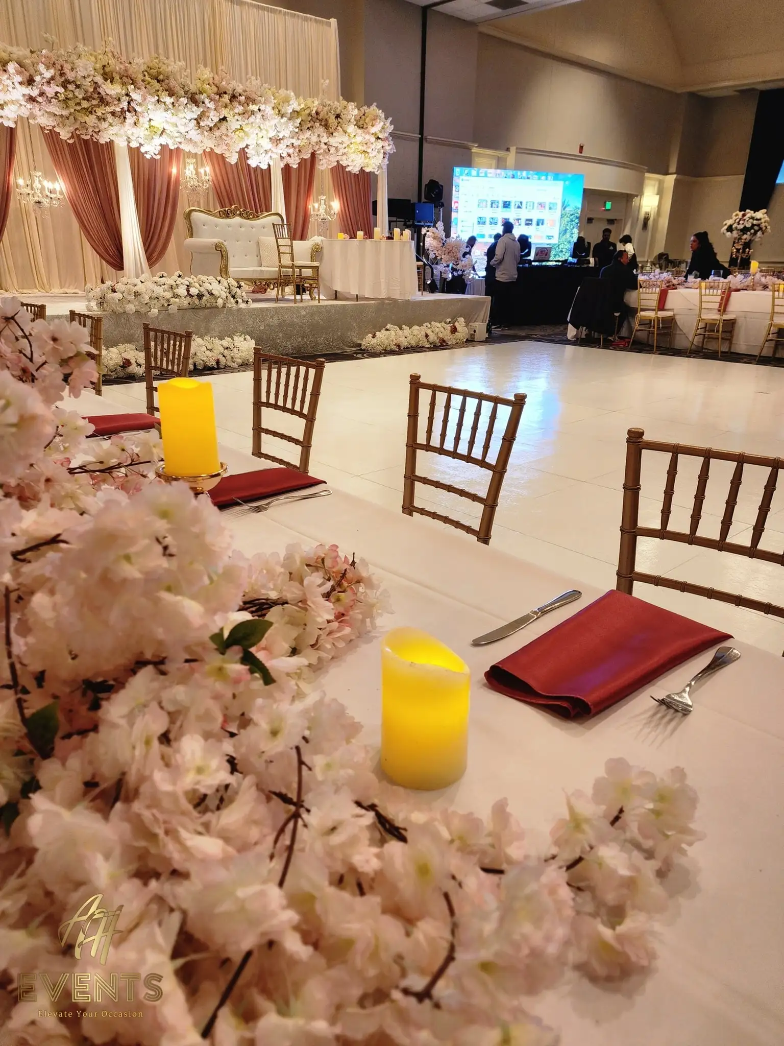 Table set with cherry-blossom branches and red napkins beside the white dance floor and floral stage