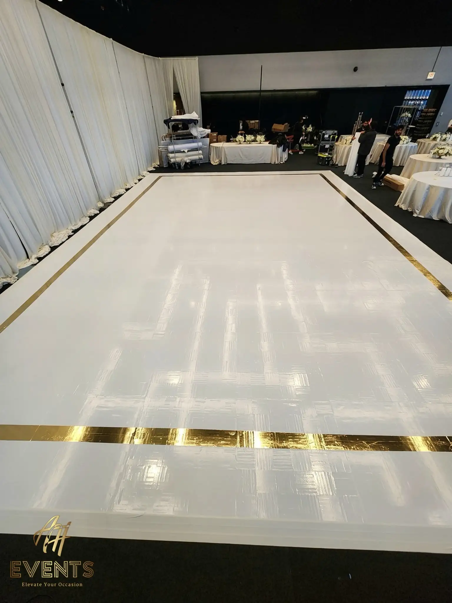 White dance floor with a gold mirror border during setup in the loft, round tables and drape behind