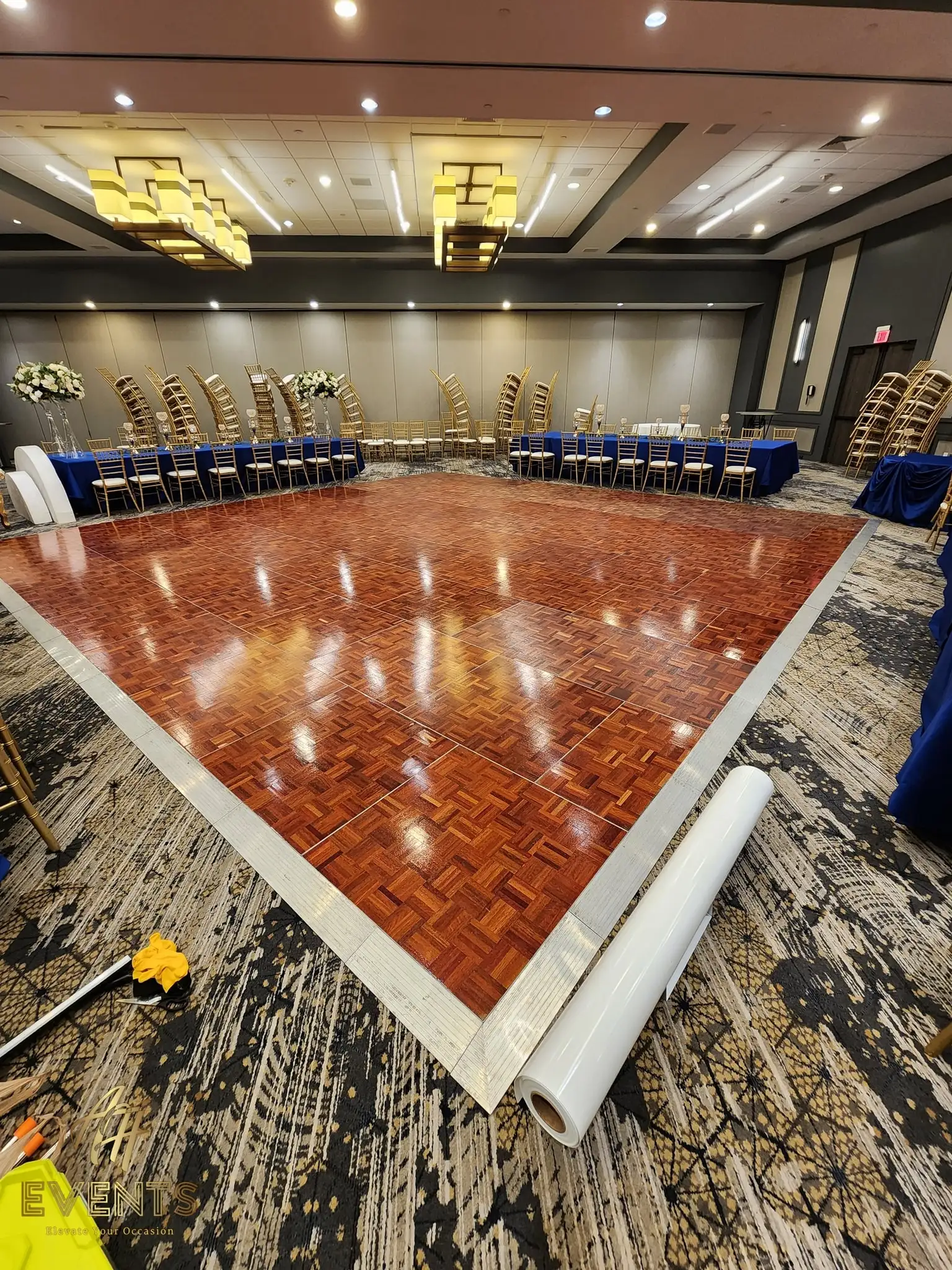 Polished parquet wood dance floor during setup, gold chiavari chairs stacked and blue linens behind