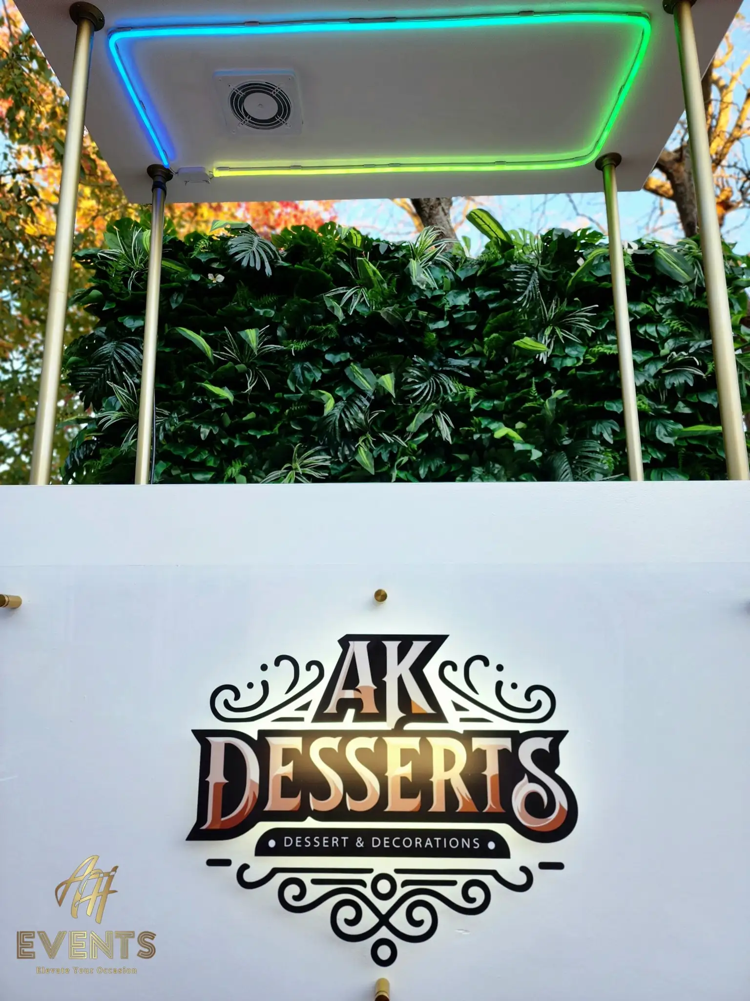 AK Desserts cart canopy lit green-and-blue with a spotlight, above the logo panel and a greenery wall