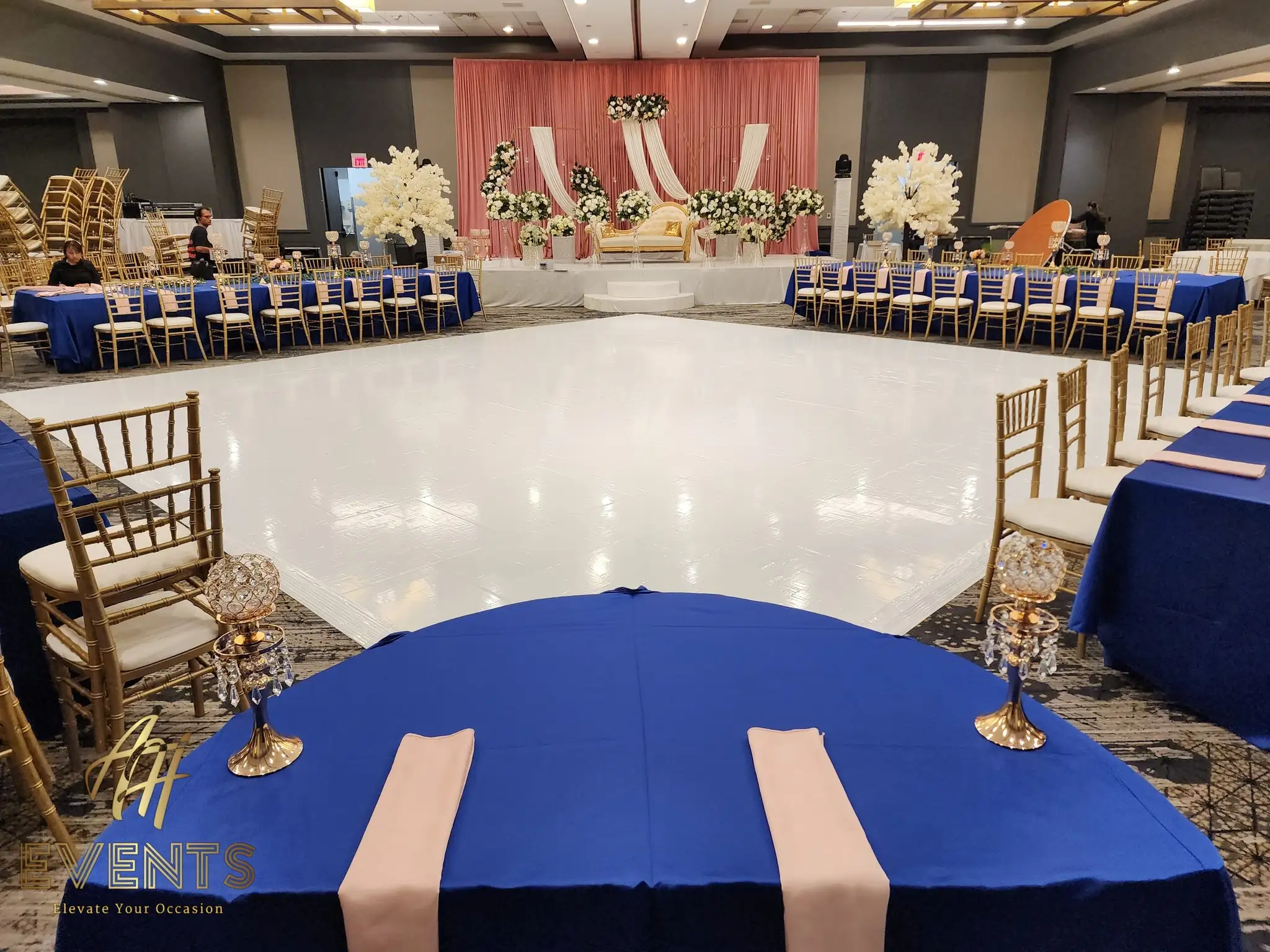 Glossy white dance floor framed by royal-blue head tables, facing a blush stage with white florals
