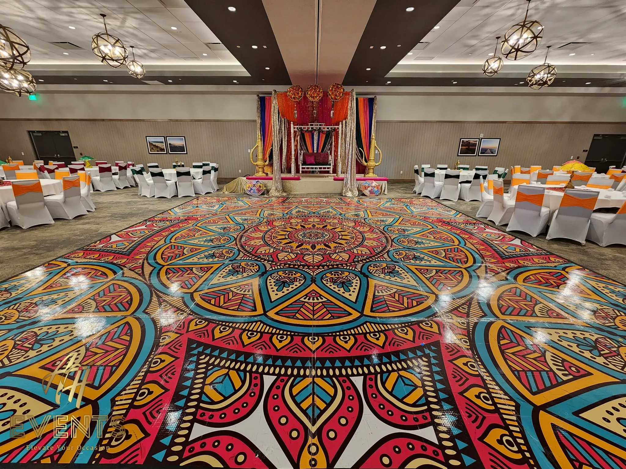 Round mandala floor under chandeliers in a banquet hall, facing a teal-draped stage with a floral swing